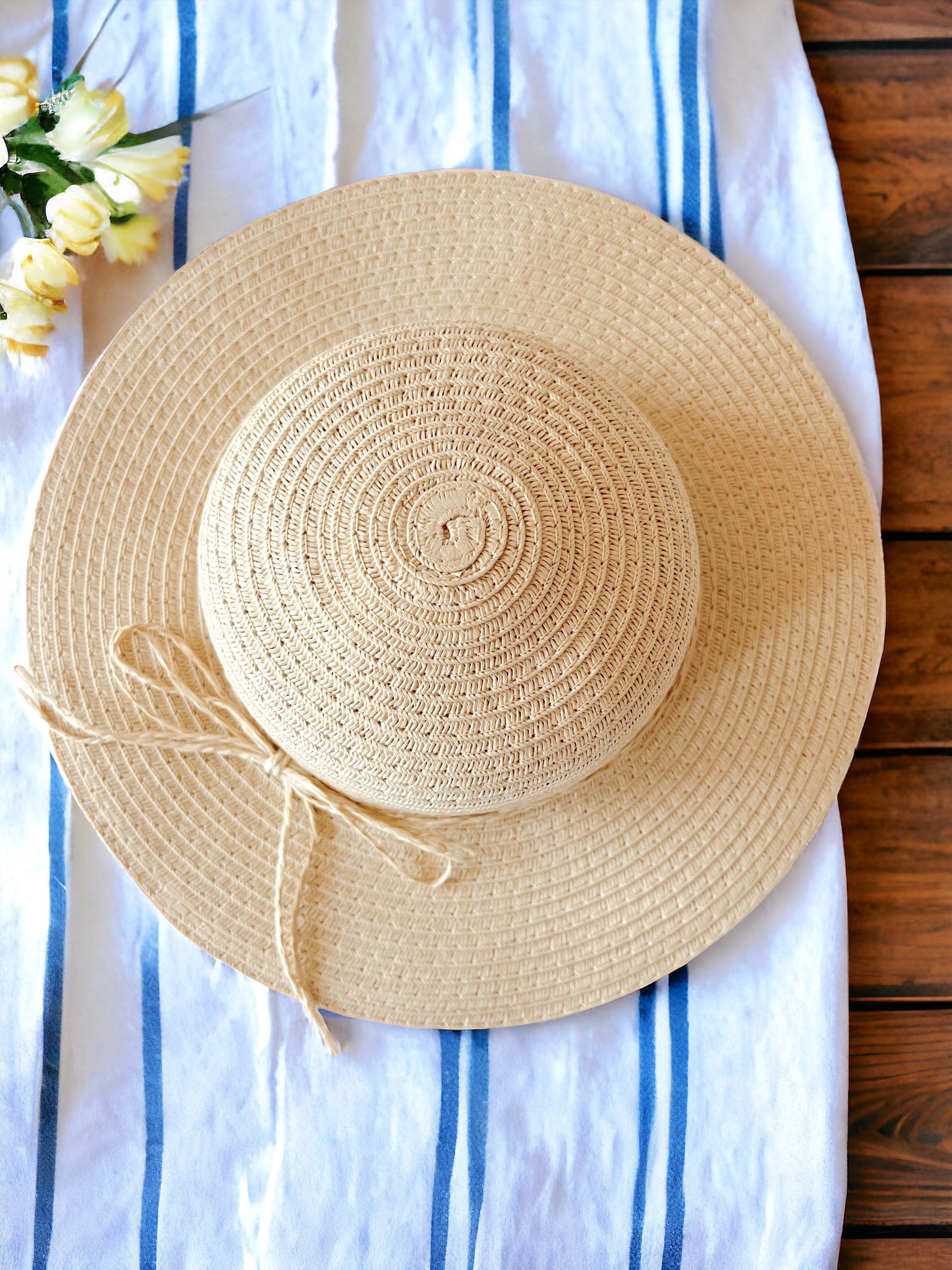 Woven straw sunhat with a ribbon, placed on a blue-striped towel and surrounded by yellow flowers.