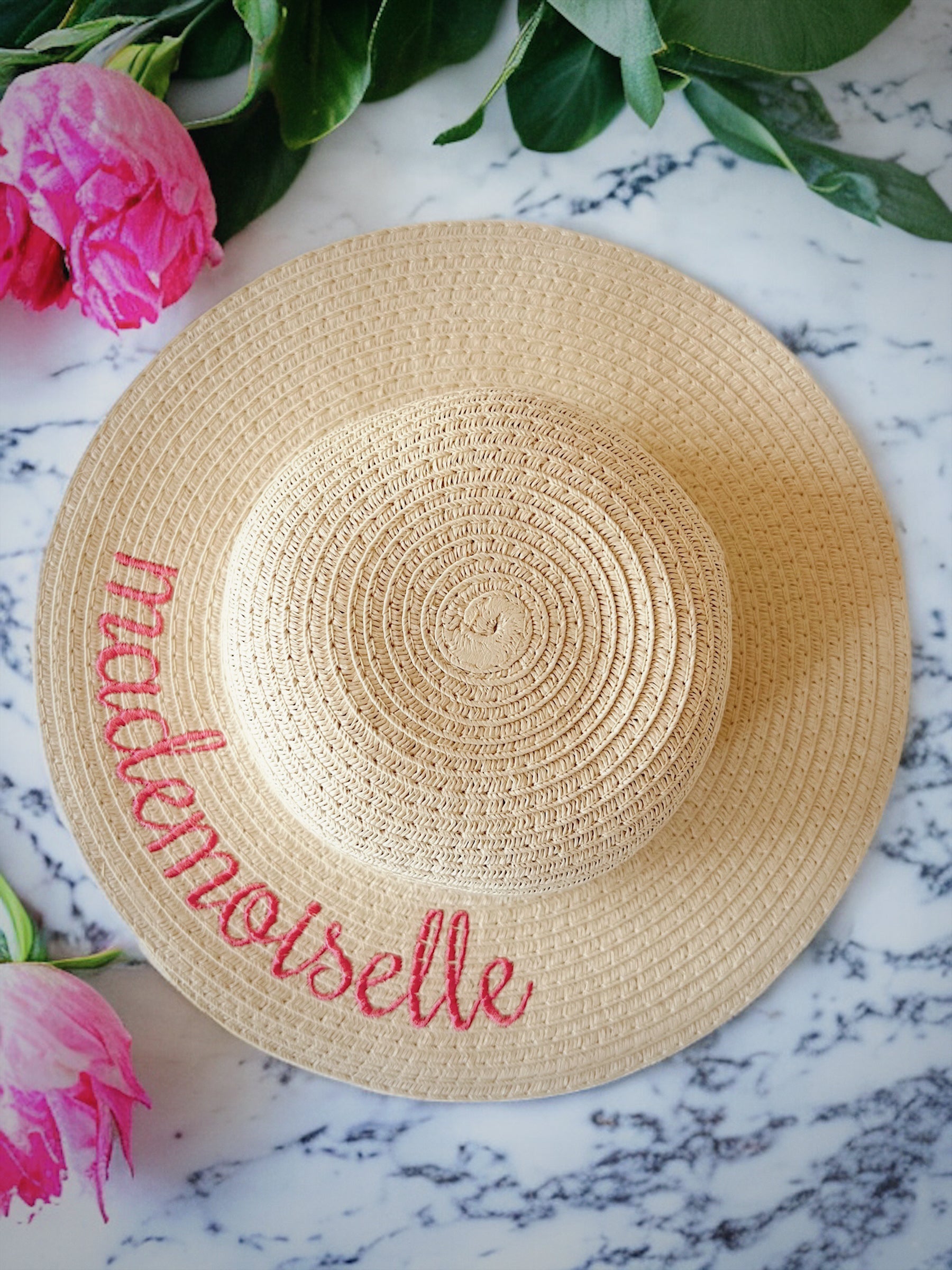 Woven straw hat with the word "mademoiselle" embroidered in red, surrounded by pink peonies and green leaves.
