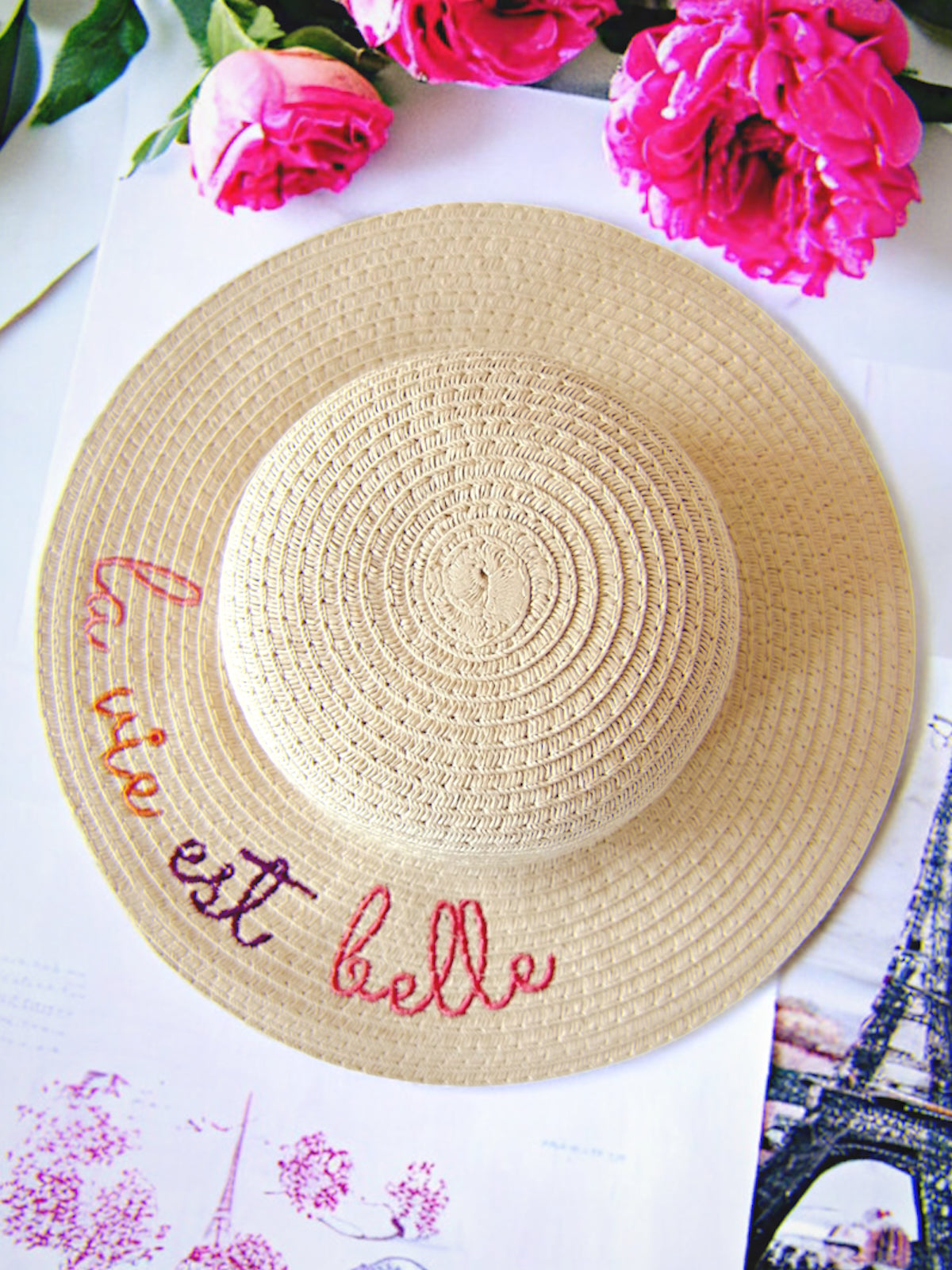 Straw hat with "la vie est belle" embroidered, surrounded by pink flowers on a white background.