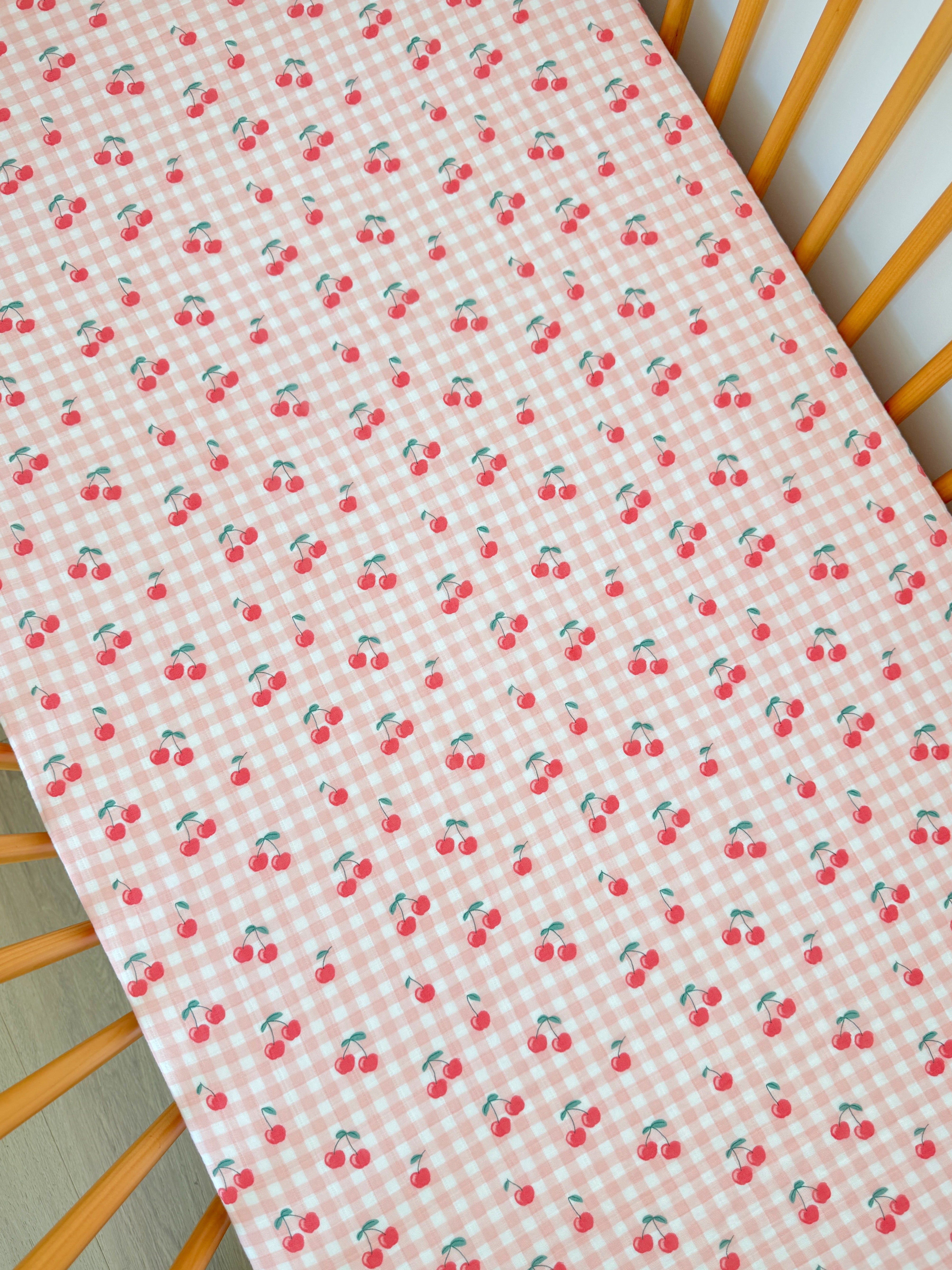 Pink checkered fabric with repeated cherry patterns, laid on a wooden crib mattress.
