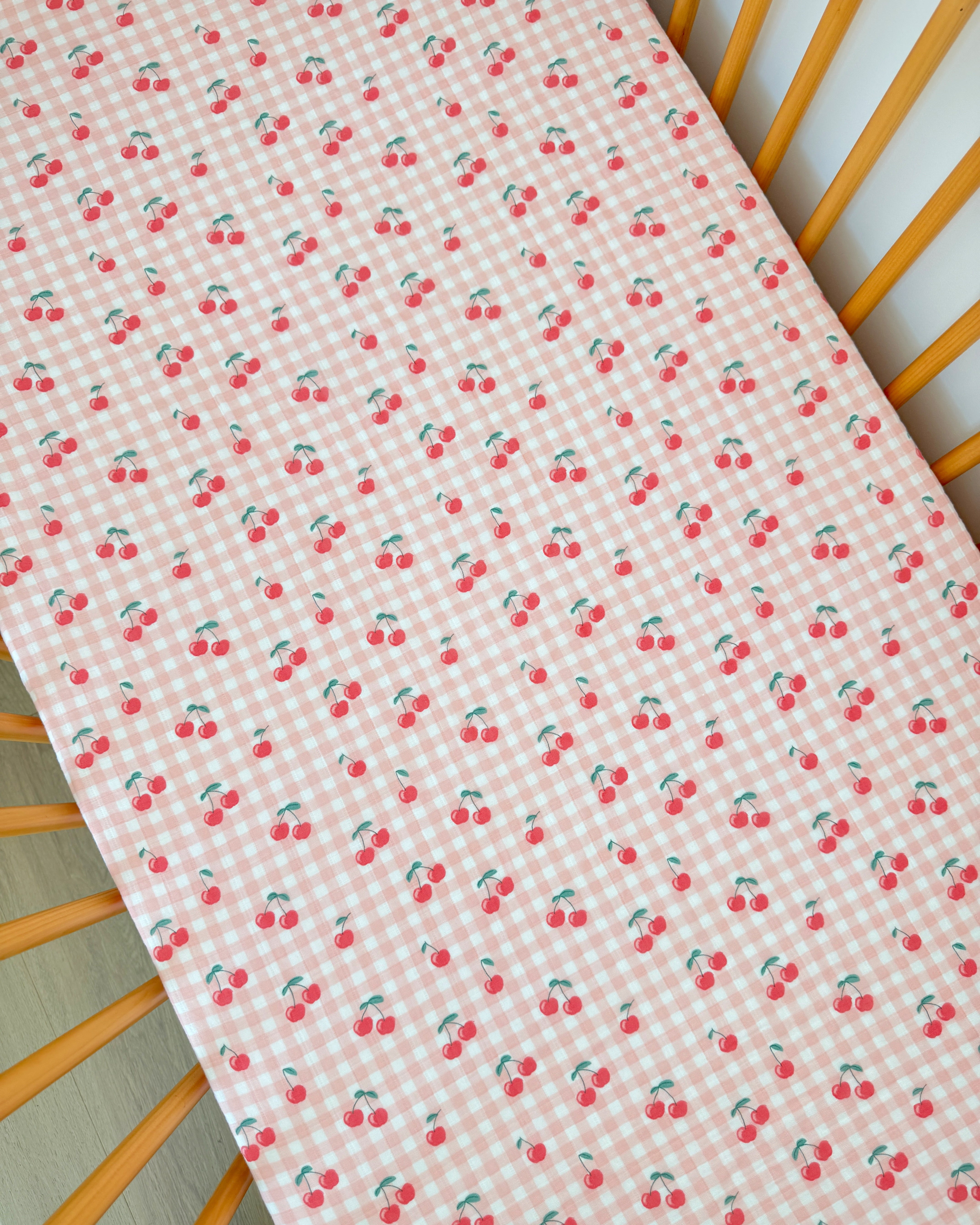 Pink checkered fabric with repeated cherry patterns, laid on a wooden crib mattress.