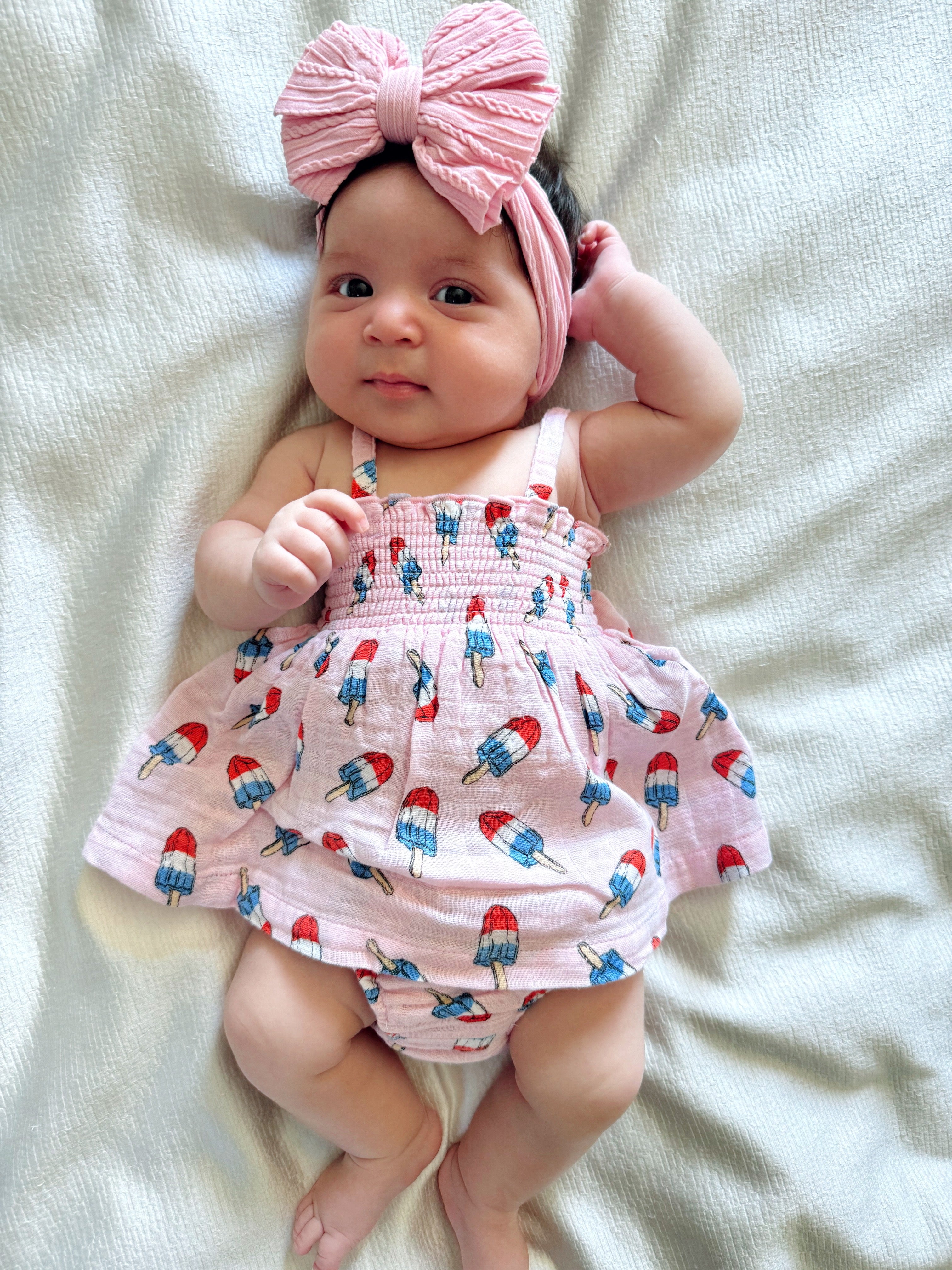 Adorable baby in a pink dress with popsicle print, wearing a large pink bow headband, lying on a soft white blanket.