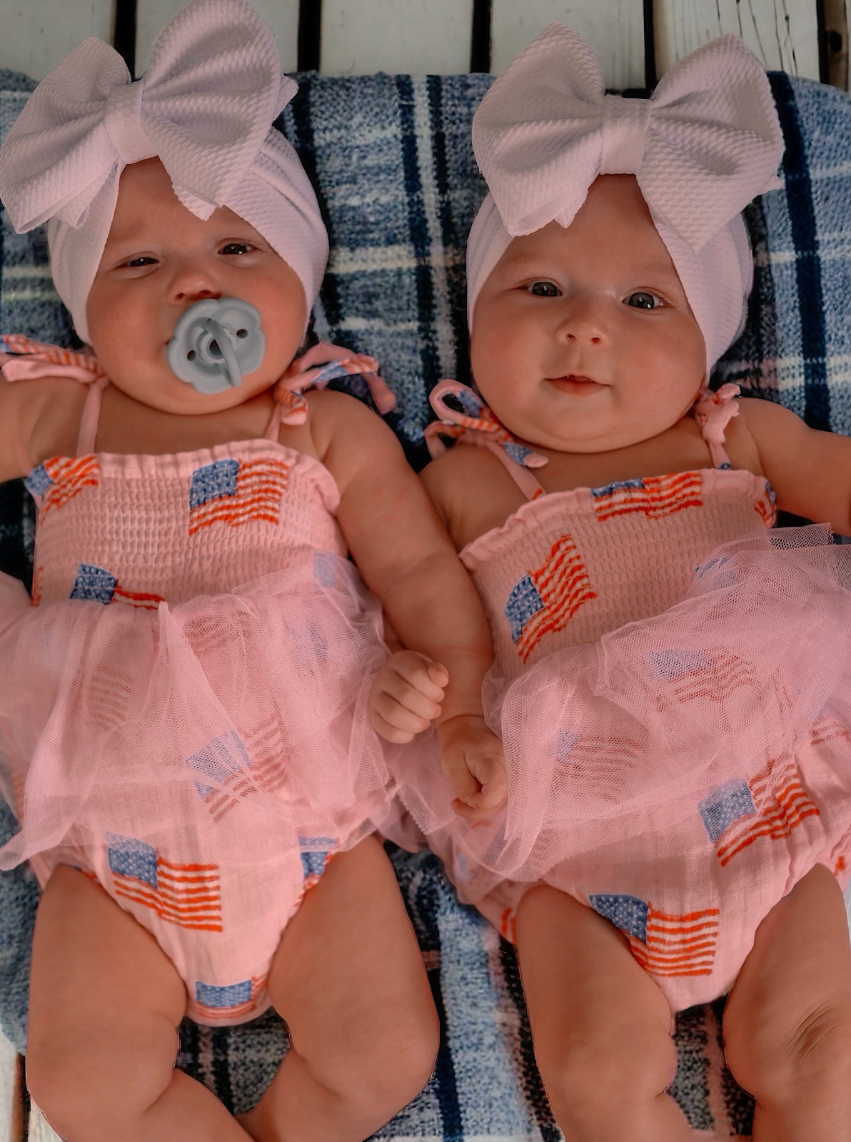 Twin babies wearing pink dresses with American flags, large bows, and pacifiers, lounging on a plaid blanket.