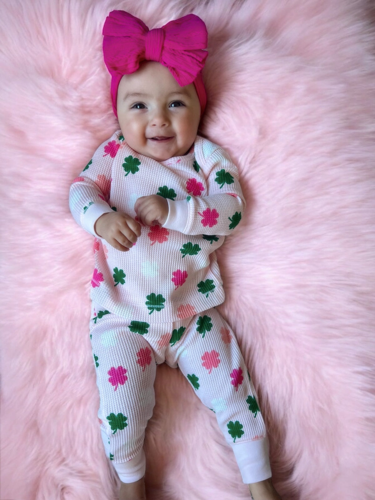 Smiling baby in a pink headband, wearing green and pink clover pajamas, lying on a fluffy pink blanket.