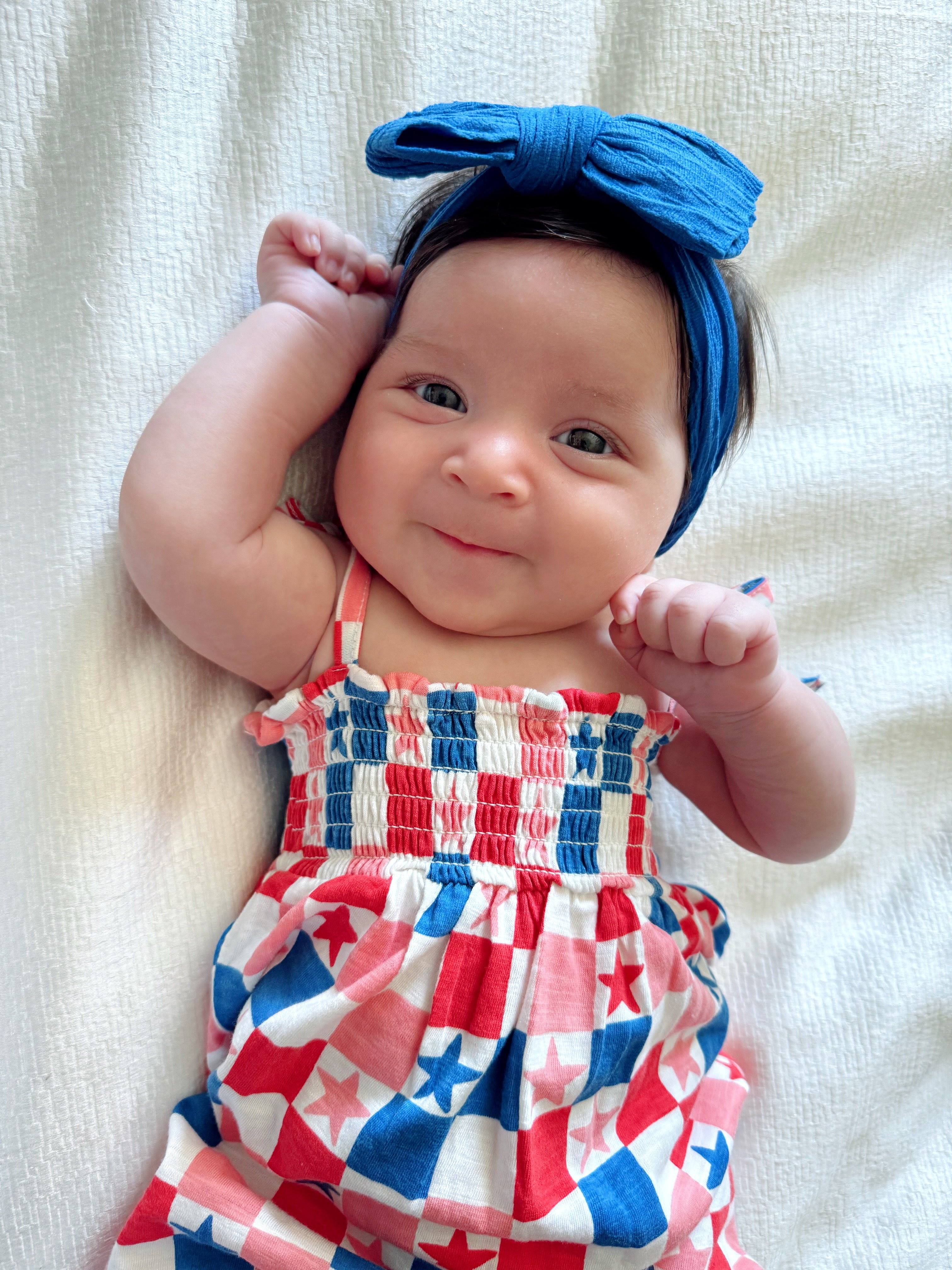 Smiling baby girl wears a blue headband and a colorful patriotic outfit on a light background.