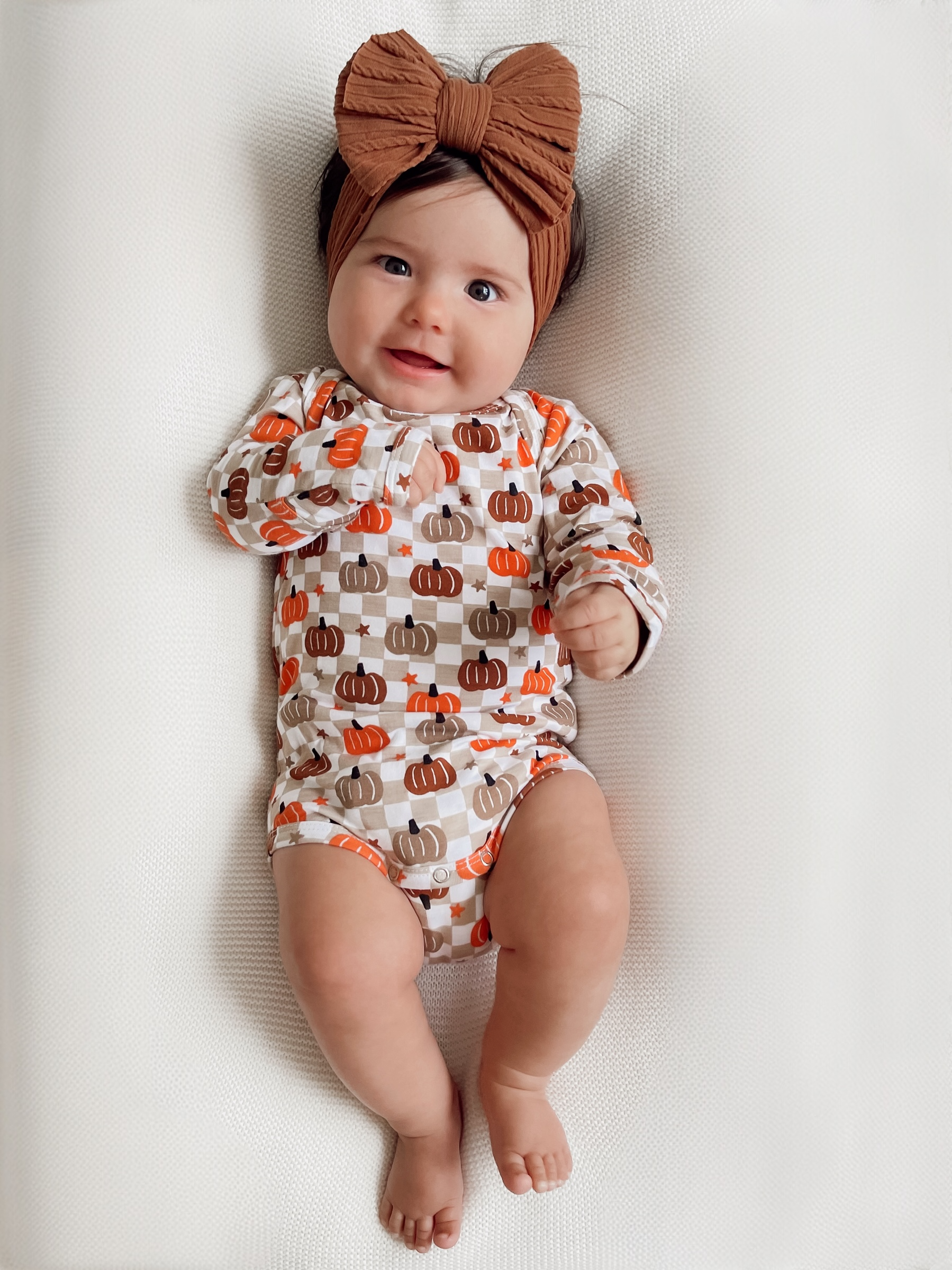 Smiling baby in a pumpkin-patterned onesie with a large brown bow, lying on a textured white surface.