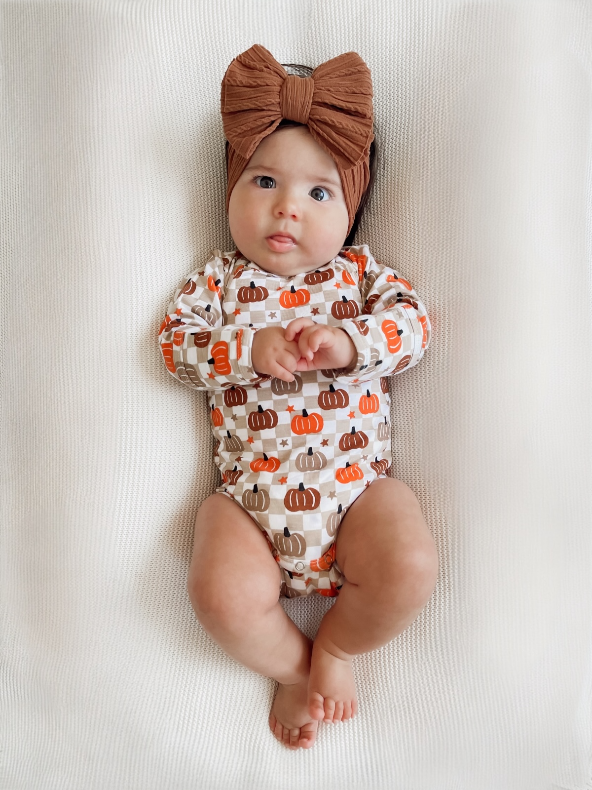 Baby wearing a pumpkin-patterned onesie and a brown bow headband, lying on a textured white blanket.