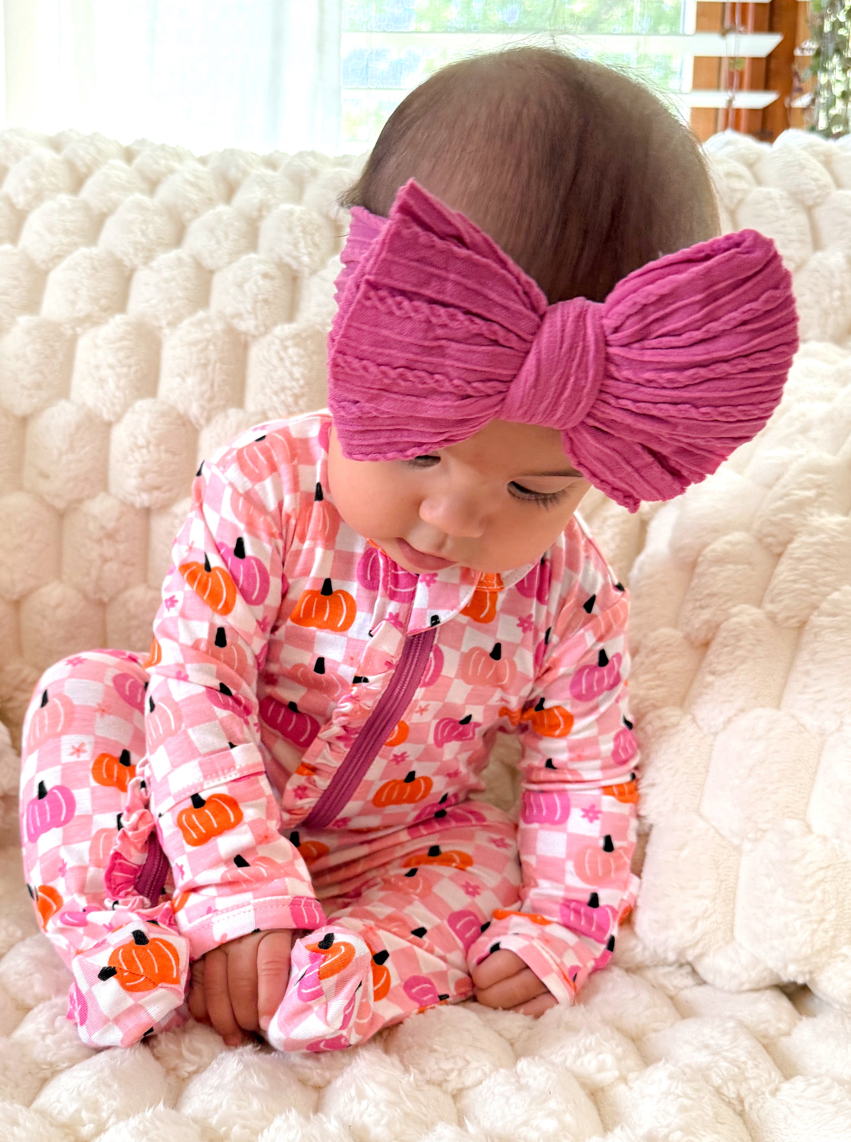 Baby in pink pumpkin-patterned outfit sits on a cozy cream blanket, wearing a large pink bow headband.