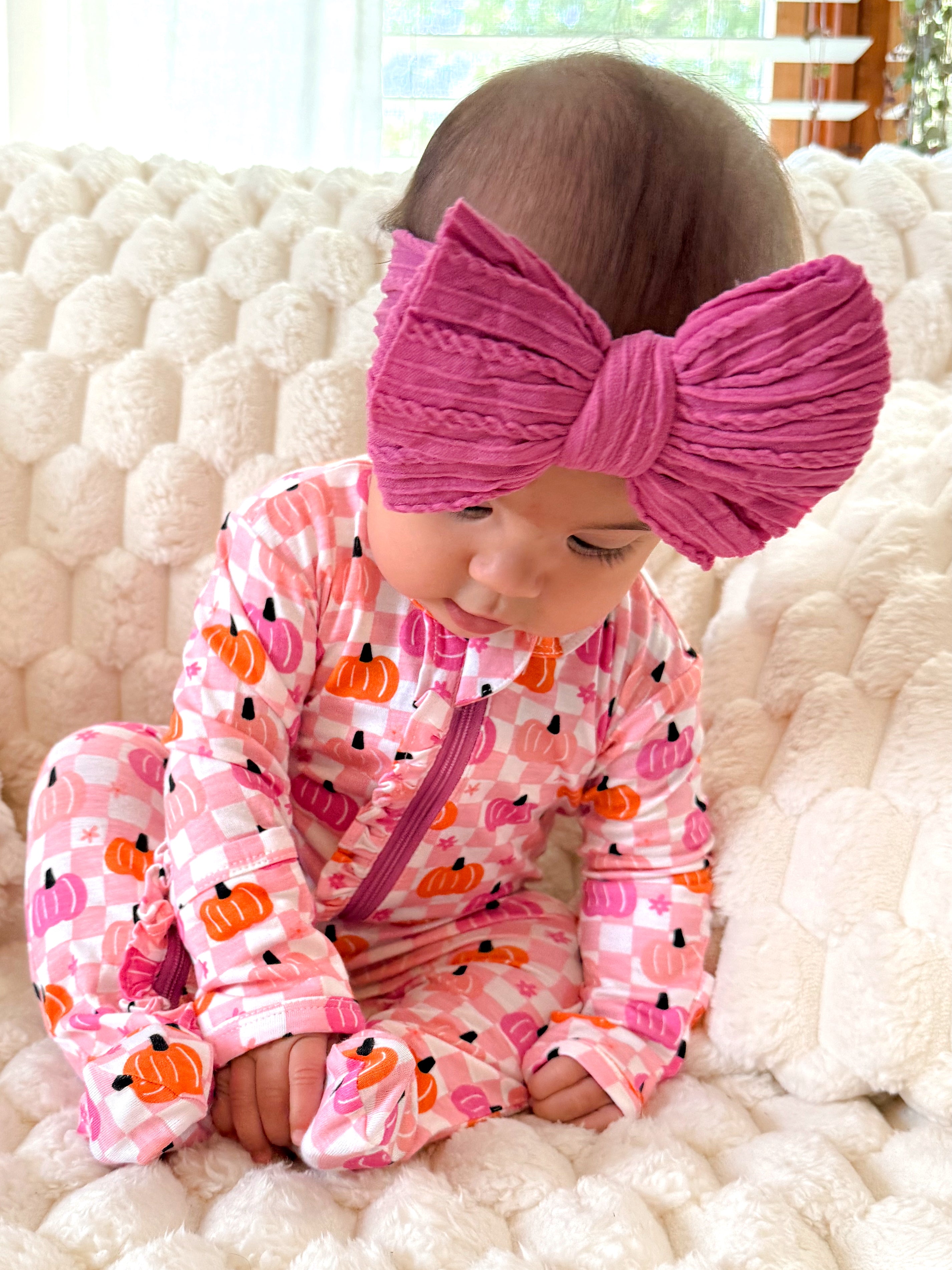 Baby in pink pumpkin-patterned outfit sits on a cozy cream blanket, wearing a large pink bow headband.
