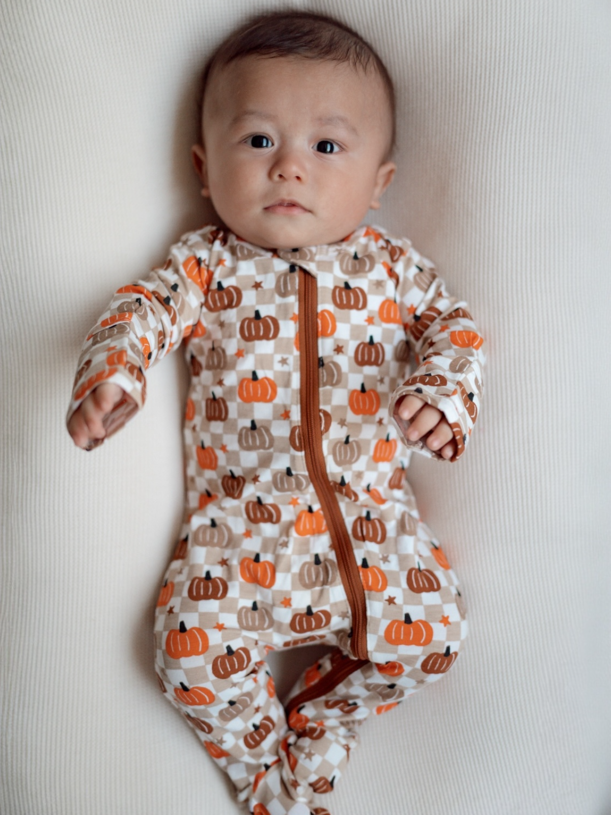 Infant wearing a pumpkin-patterned onesie, lying on a light textured surface, with a neutral expression.