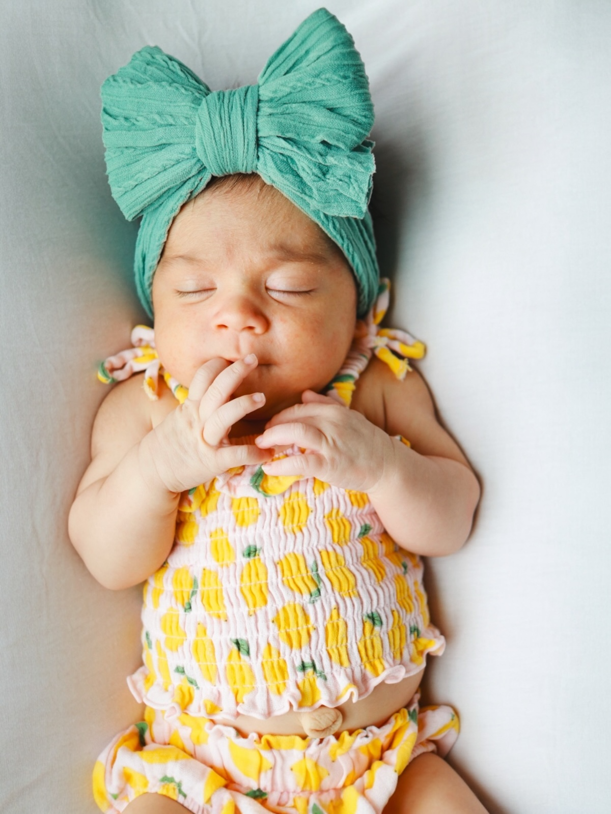 Sleeping baby with a green bow headband, wearing a colorful, patterned outfit, resting on a light background.