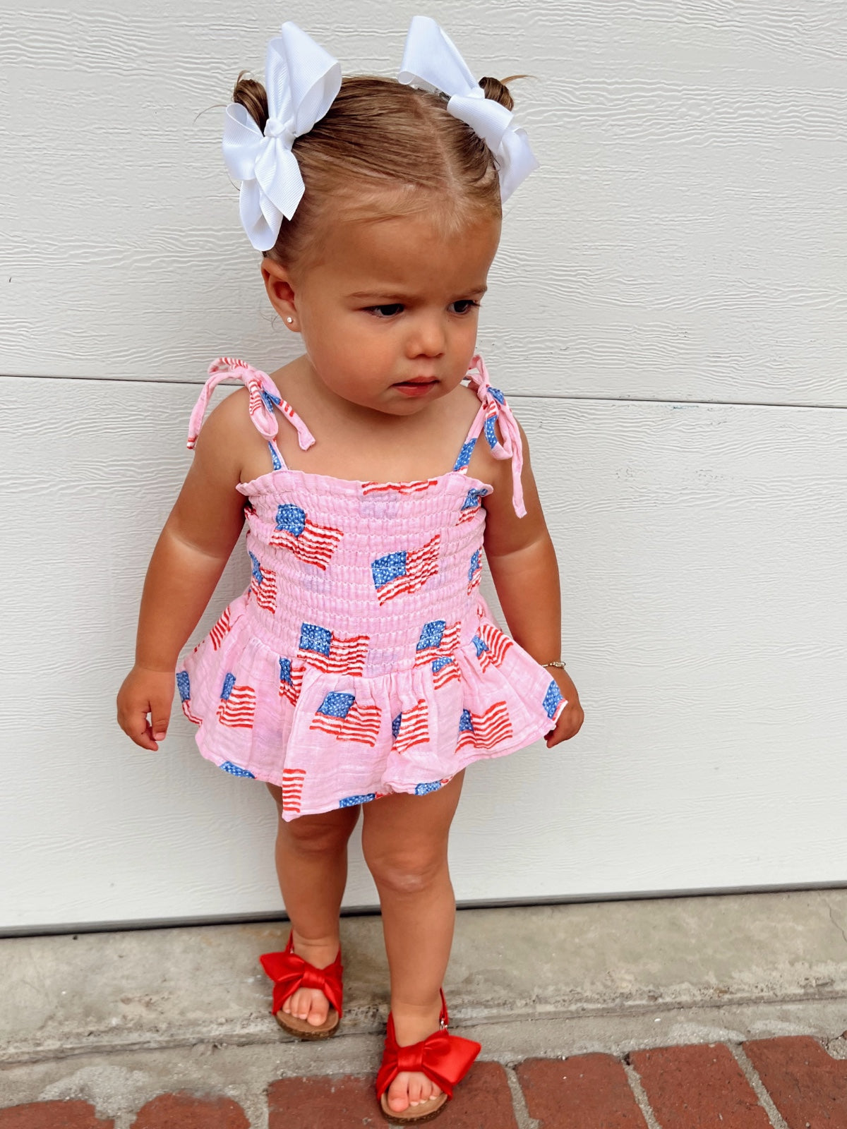 Toddler in a patriotic dress with American flags, white bows in hair, standing against a light background.