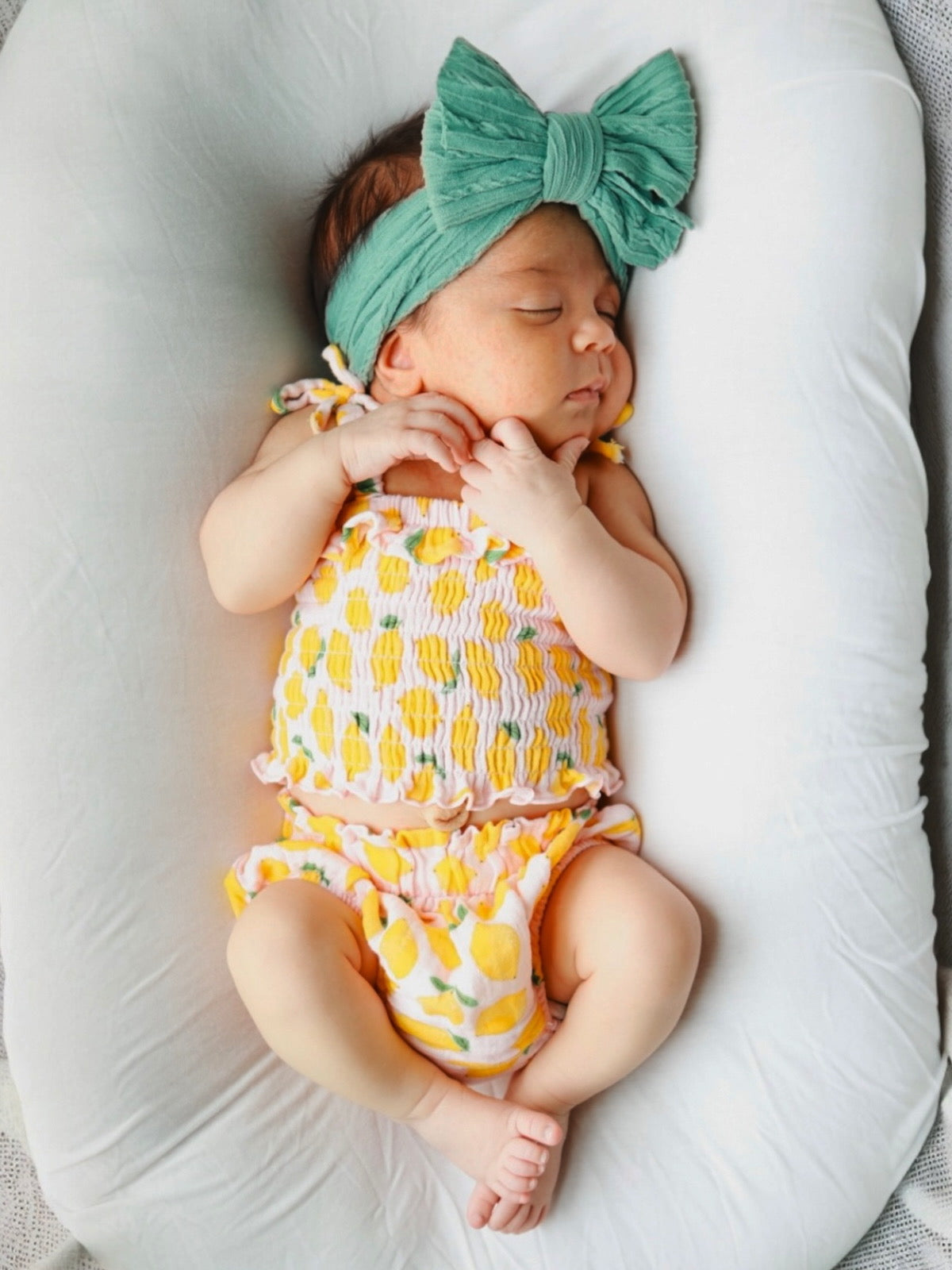 Sleeping baby girl with a green bow headband, wearing a colorful, frilly outfit, resting on a soft pillow.