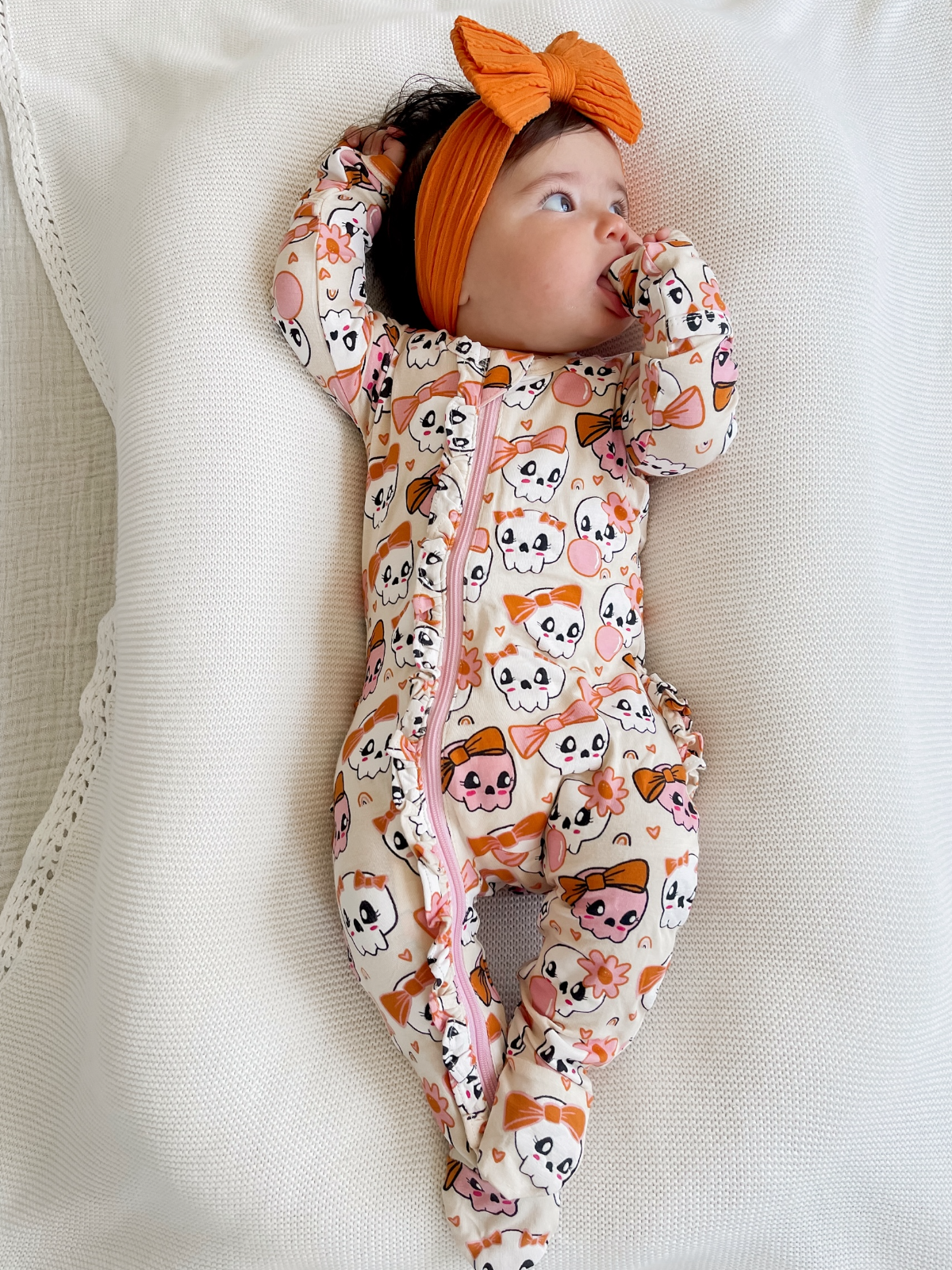 Infant in colorful skull-print pajamas and an orange headband, lying on a textured white blanket.