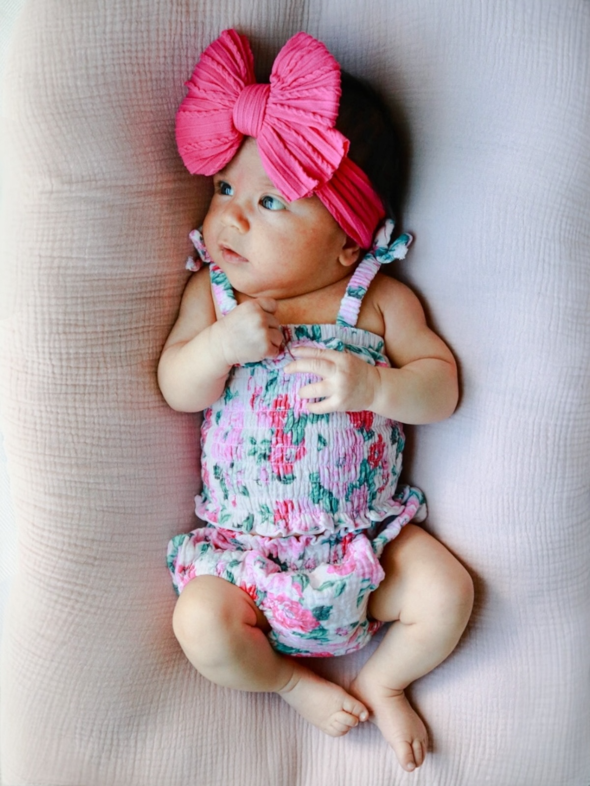 Baby girl wearing a floral romper and a large pink bow headband, lying on a soft pink blanket.