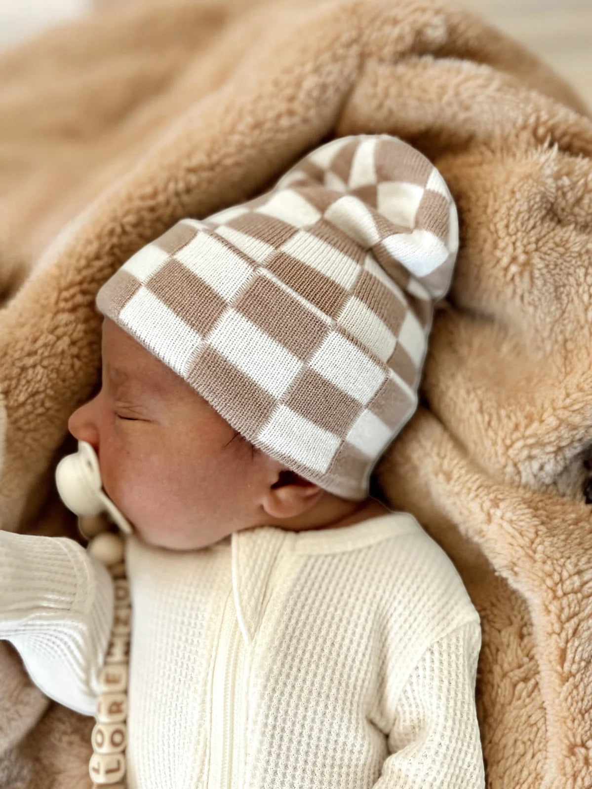 Baby in a checkered beanie, sleeping on a soft blanket, with a pacifier nearby.