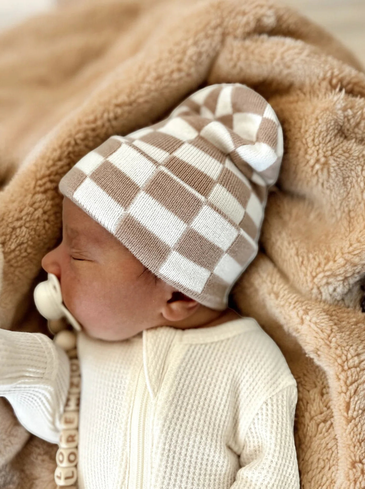 Baby in a checkered beanie, sleeping on a soft blanket, with a pacifier nearby.