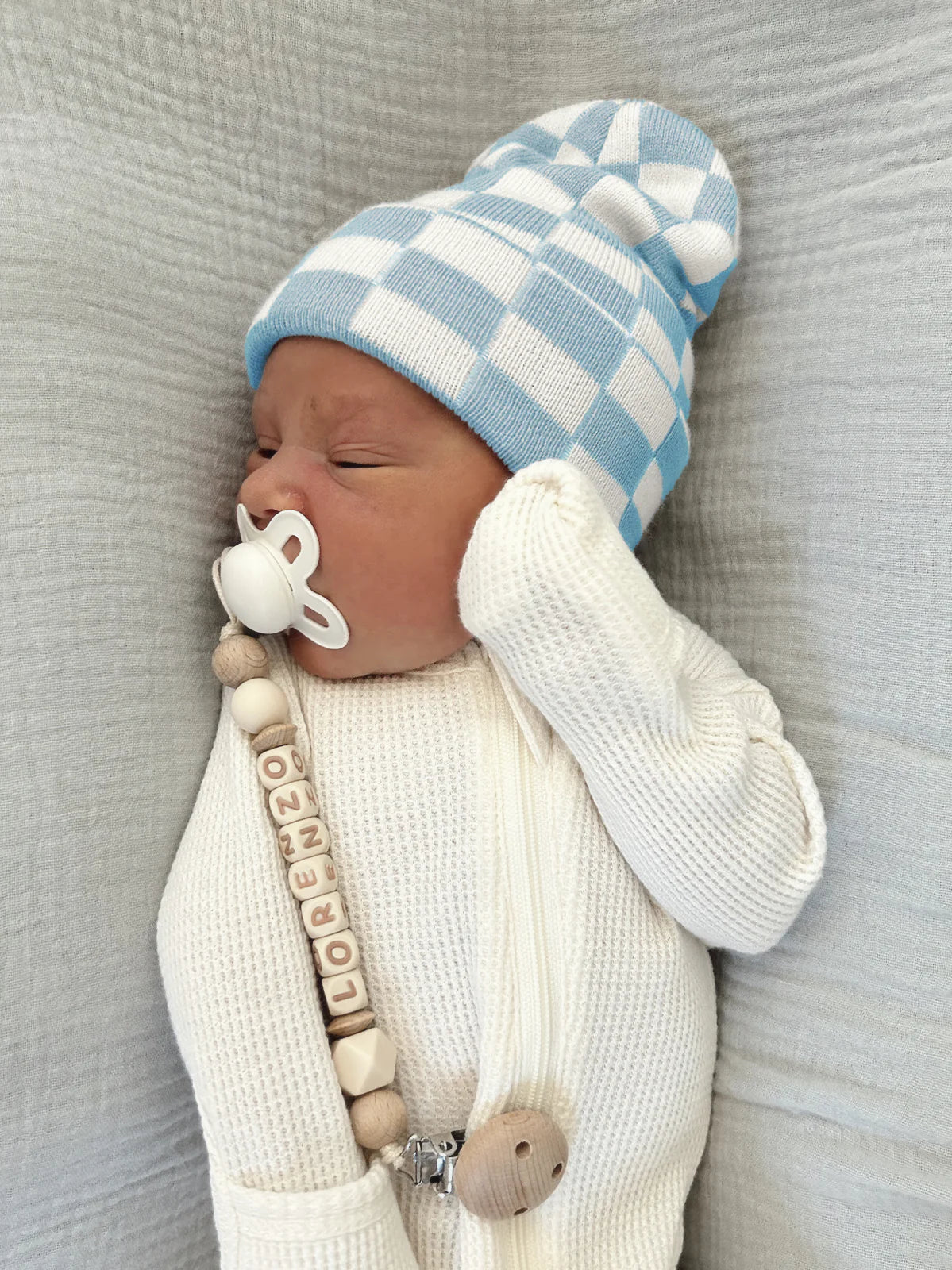 Newborn baby in a blue and white checkered hat, wearing a cream sweater with a pacifier and a name bead clip.