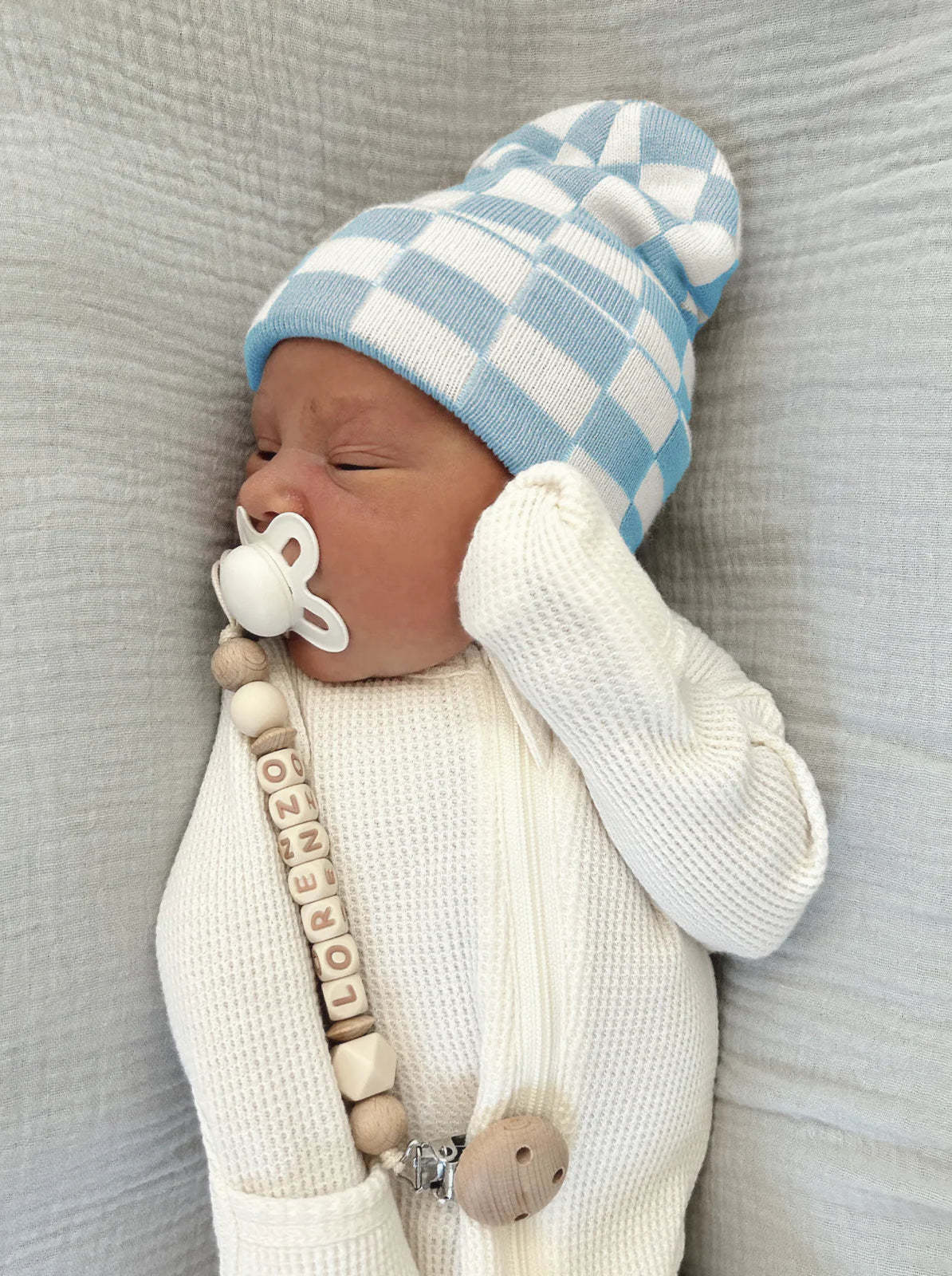 Newborn baby in a blue and white checkered hat, wearing a cream sweater with a pacifier and a name bead clip.
