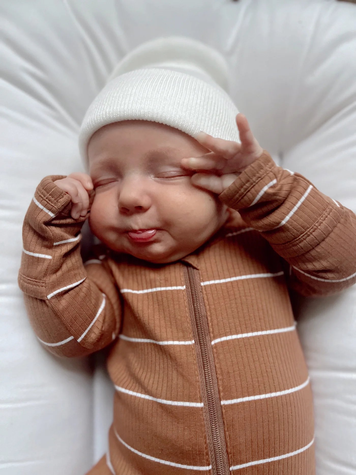 Baby with a playful expression, wearing a brown striped onesie and white beanie, sticking out their tongue.