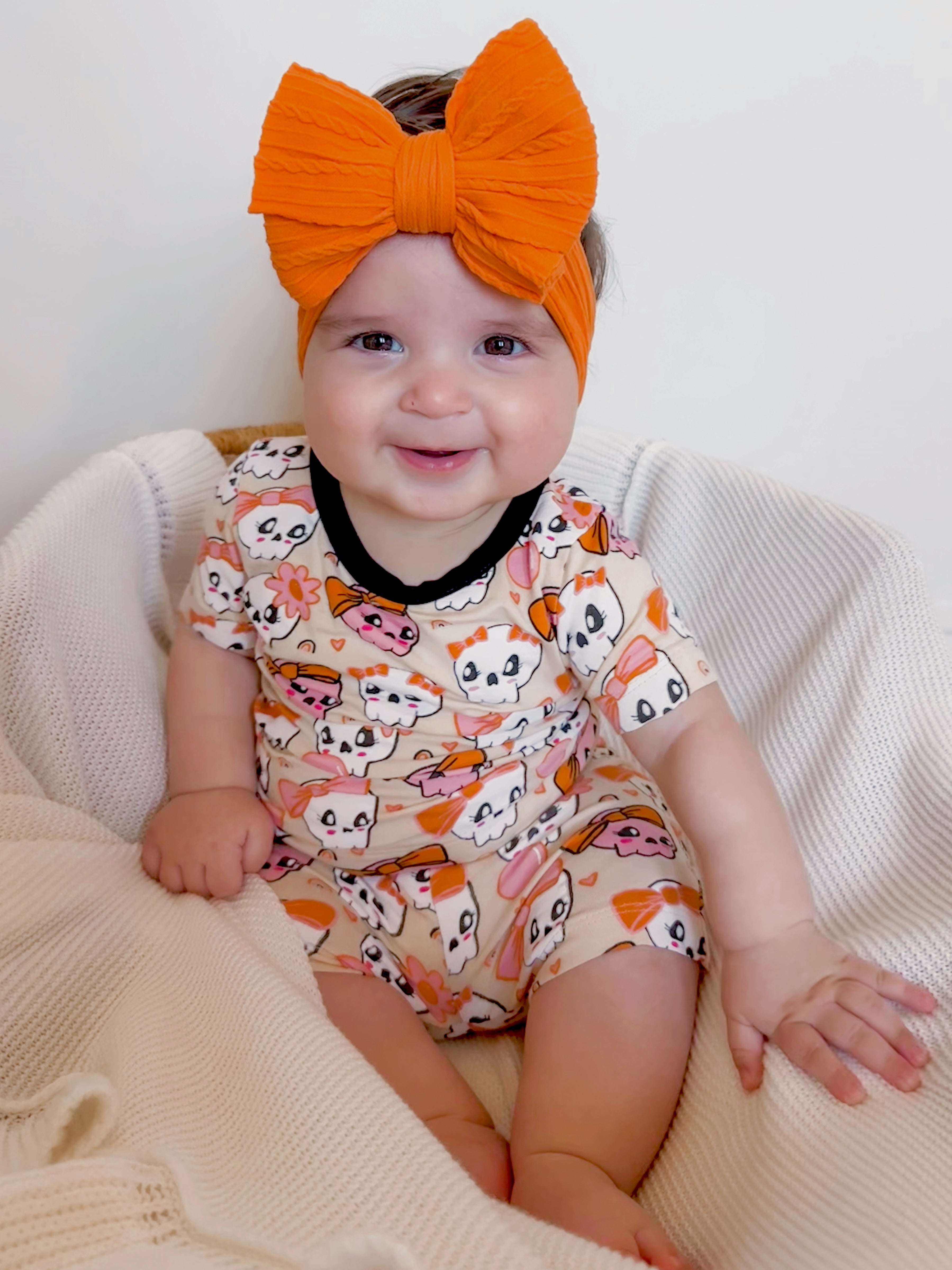 Smiling baby in a cute animal print onesie and large orange headband, sitting on a cozy blanket.