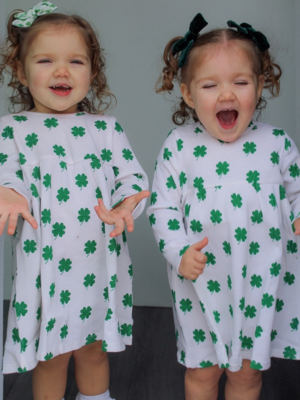Two smiling toddlers in matching white dresses with green shamrocks, posing playfully with hands outstretched.