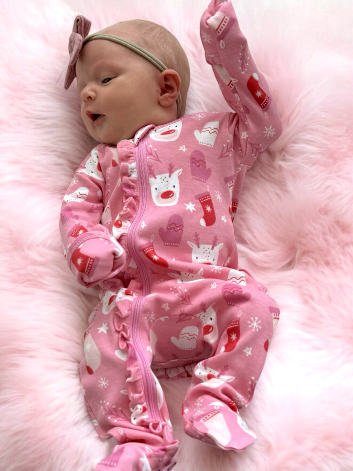 Infant in pink festive onesie with reindeer and holiday motifs, lying on a soft pink fur blanket.