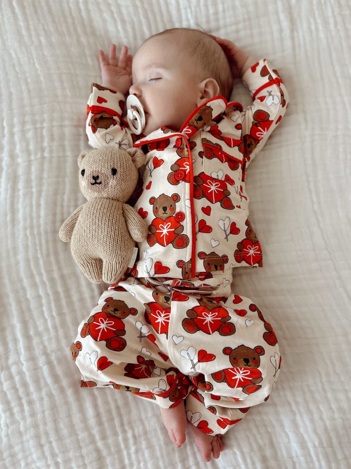 Baby sleeping in bear-patterned pajamas with a stuffed bear, peacefully resting on a white quilt.