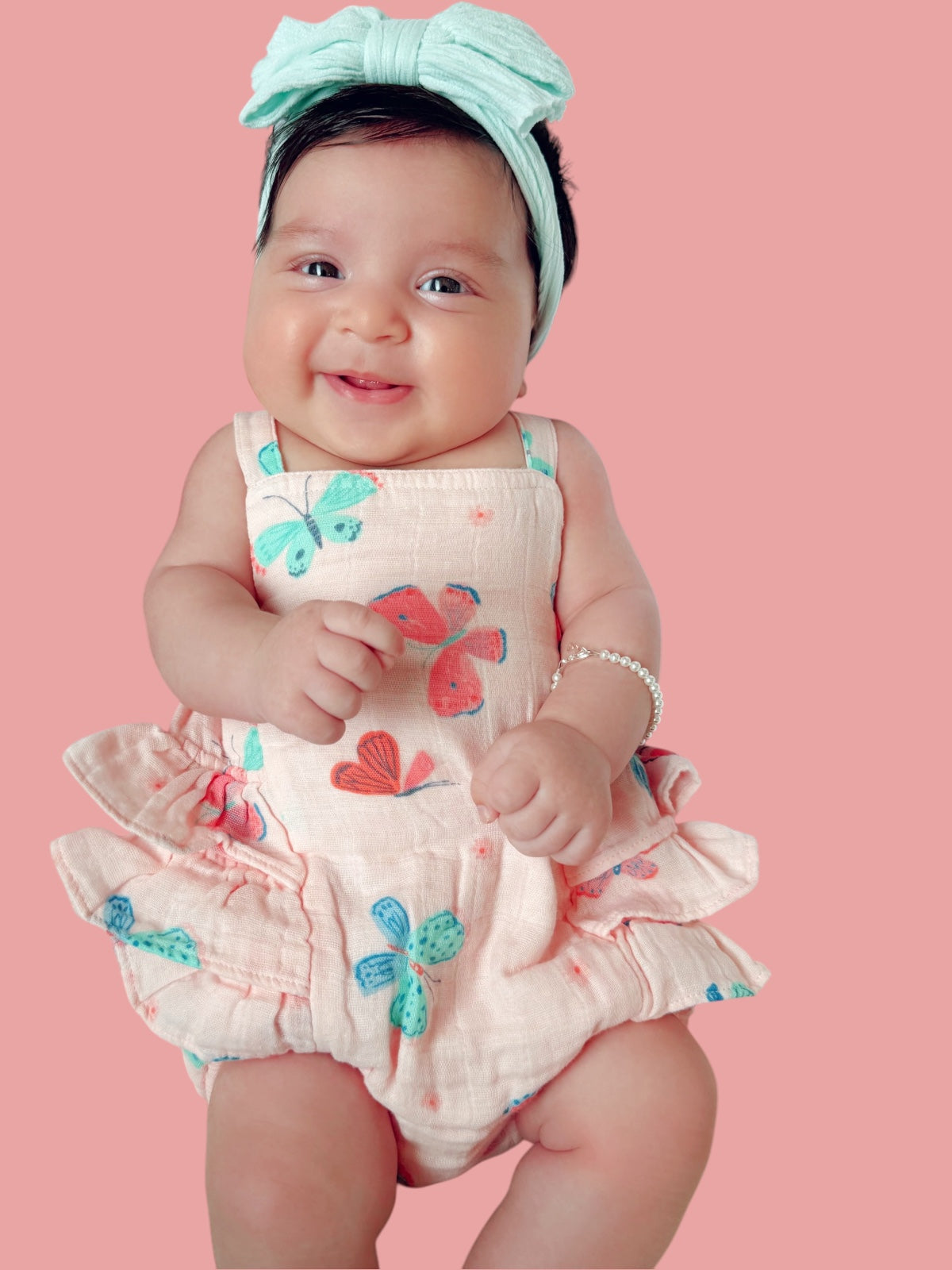 Smiling baby girl wearing a pink floral outfit and a mint green headband against a pink background.