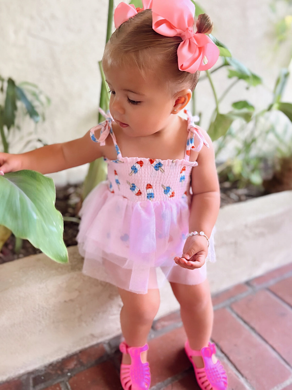 Toddler in a floral smocked dress and pink sandals, exploring a green plant in a sunny outdoor setting.