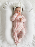 Smiling baby in pink gingham pajamas and white bow, lying on a textured blanket.