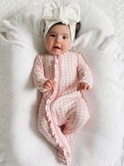 Baby in pink gingham onesie with ruffles and a white bow headband, lying on a soft textured blanket.