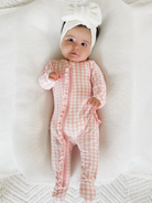 Baby girl in pink gingham romper and white bow, lying on textured white blanket, smiling at the camera.