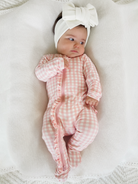 Baby girl in pink gingham pajamas and a large bow headband, resting on a textured white blanket.