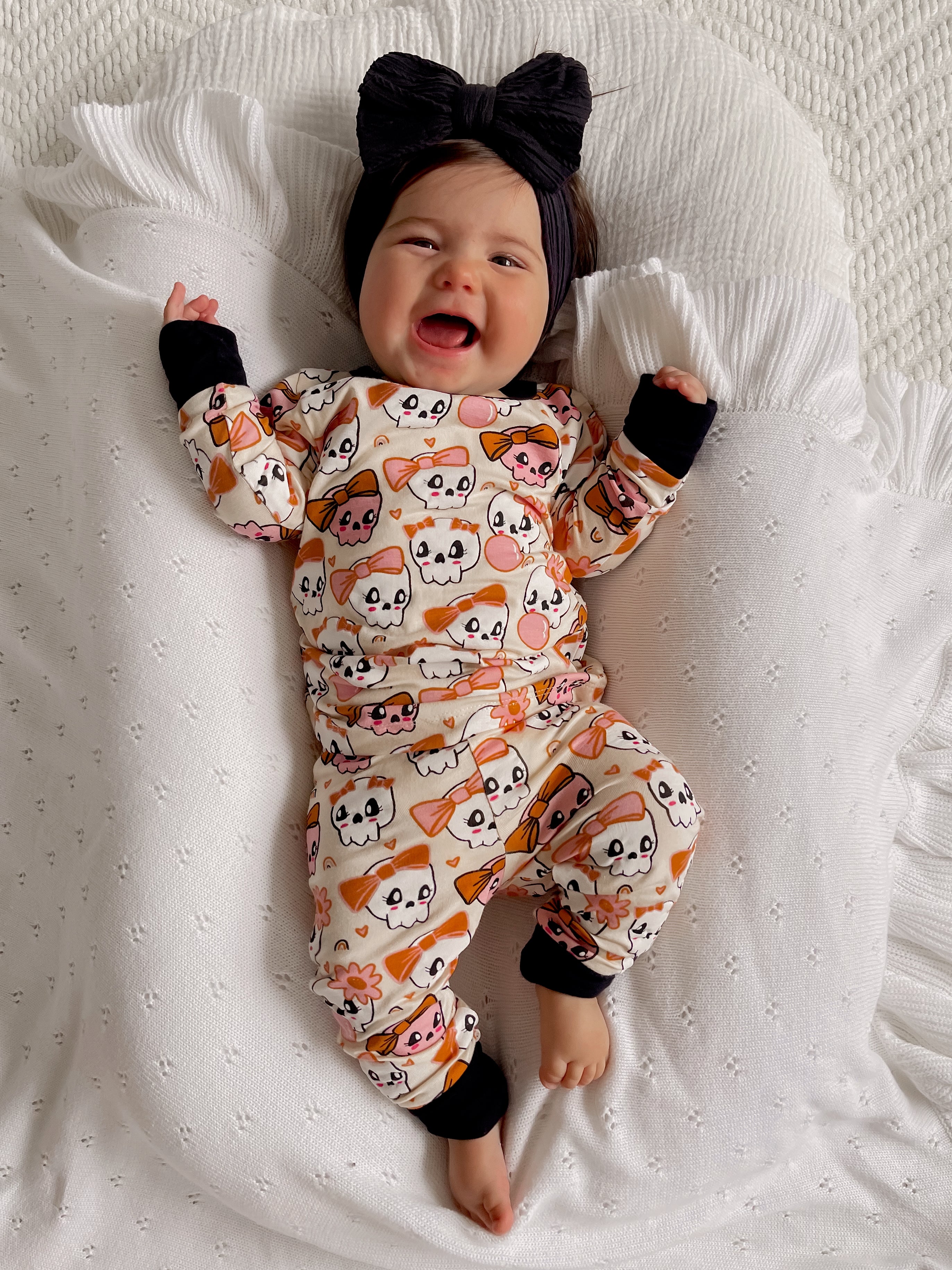 Smiling baby in cute ghost-patterned pajamas, wearing a large black bow, on a cozy blanket.