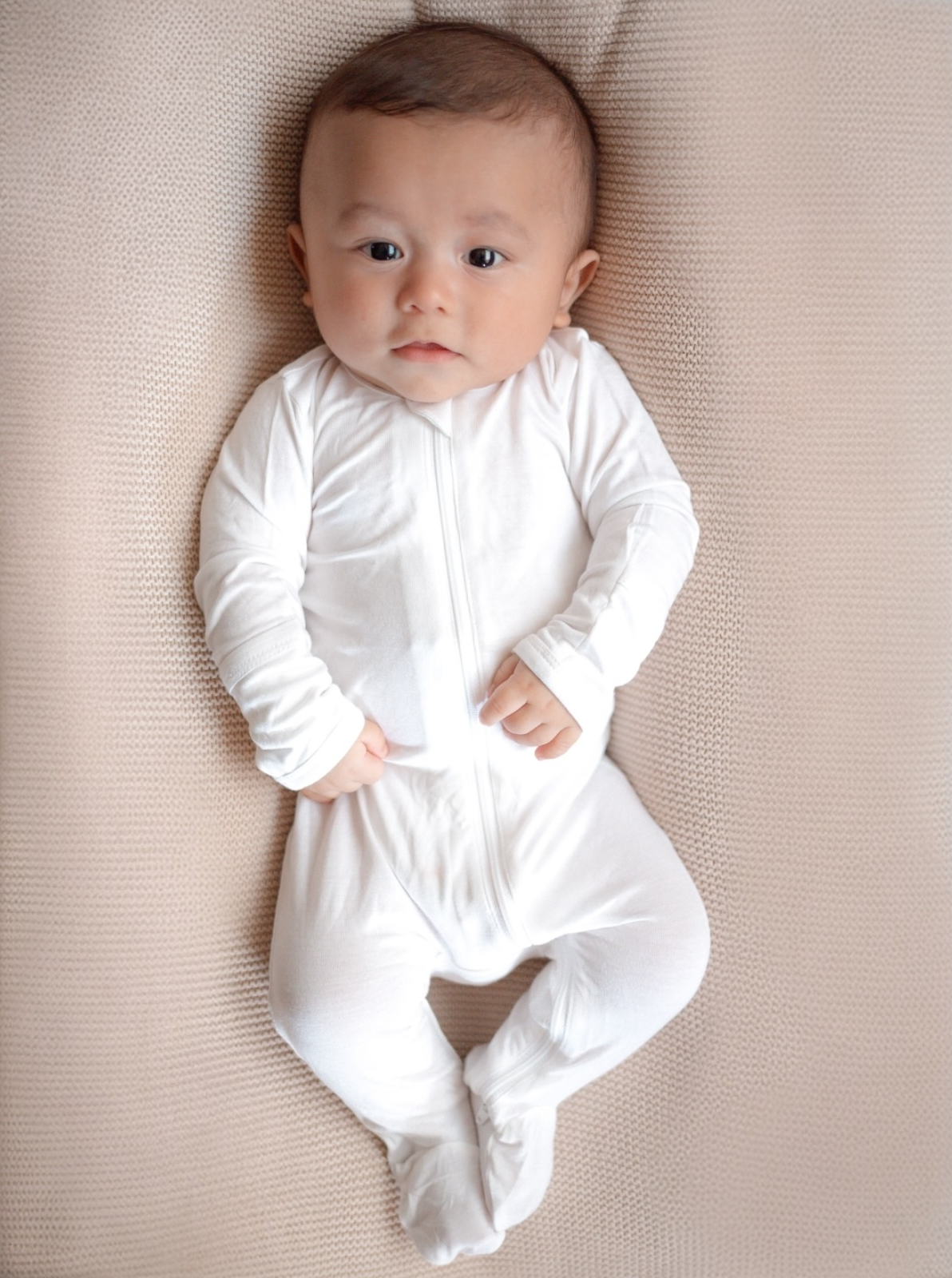 Baby in a white onesie lying on a soft, textured blanket, looking directly at the camera.