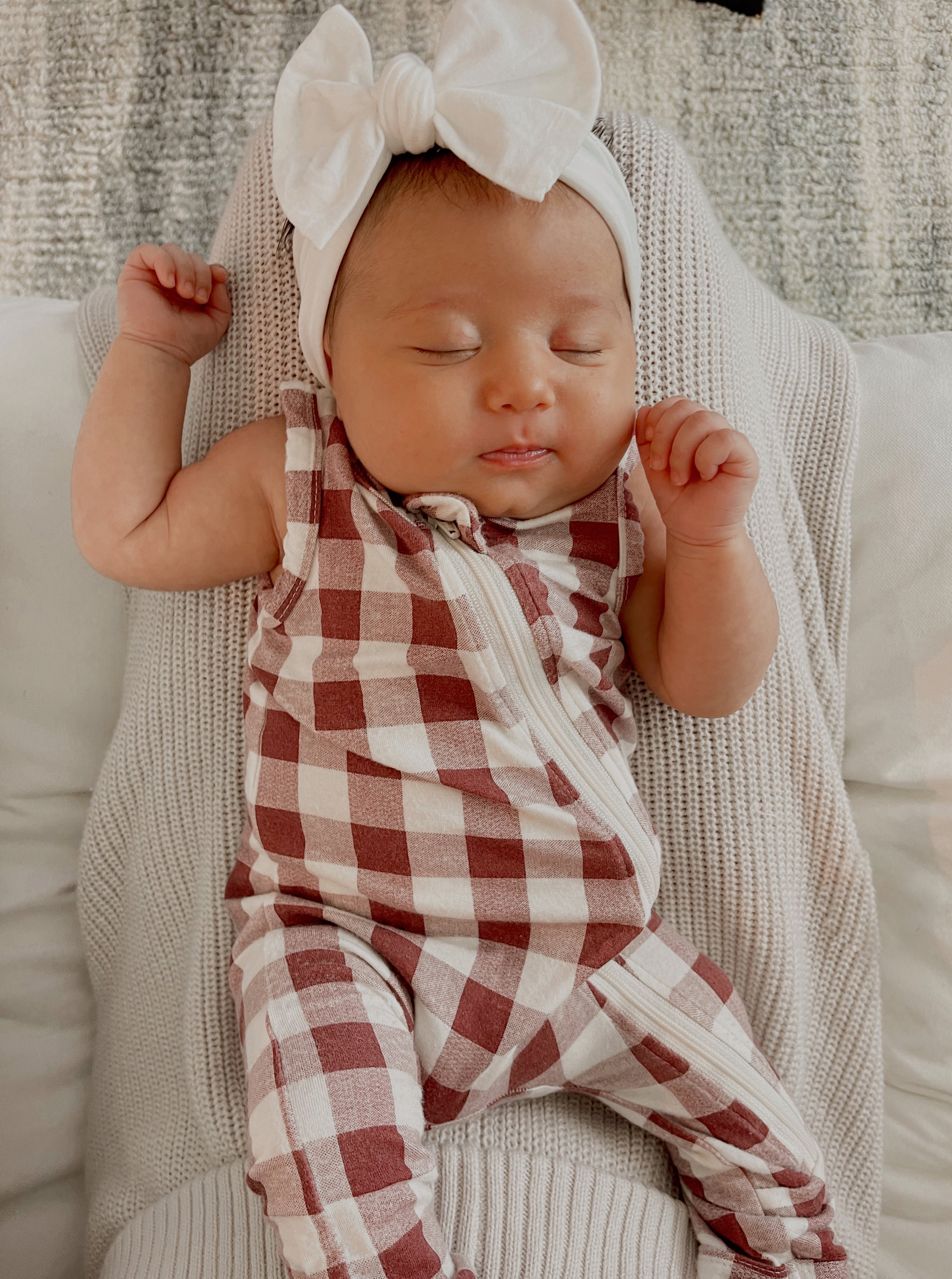 Baby girl sleeping on a white blanket, dressed in a red and white checkered onesie with a large white bow headband.