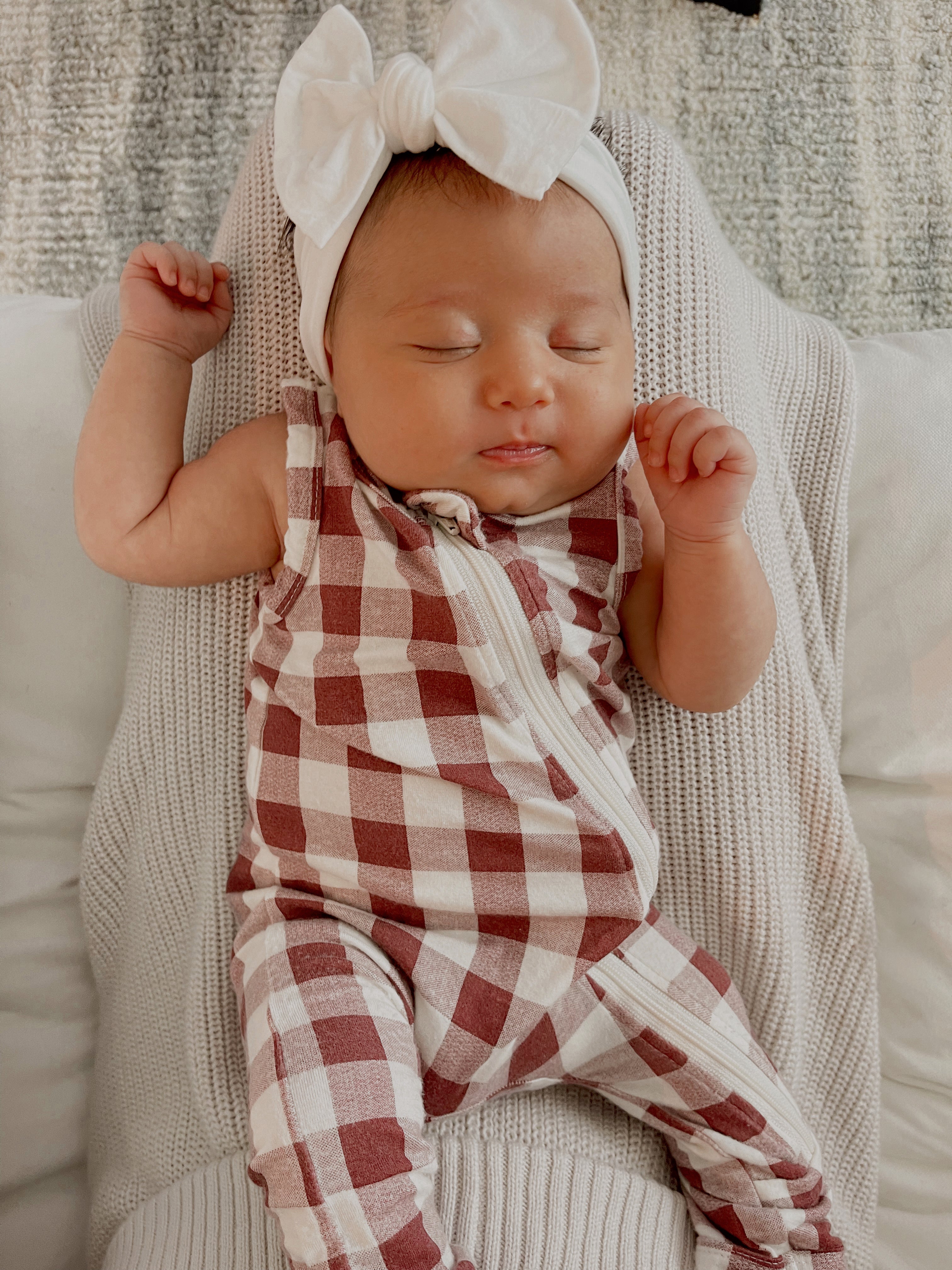 Baby girl sleeping on a white blanket, dressed in a red and white checkered onesie with a large white bow headband.