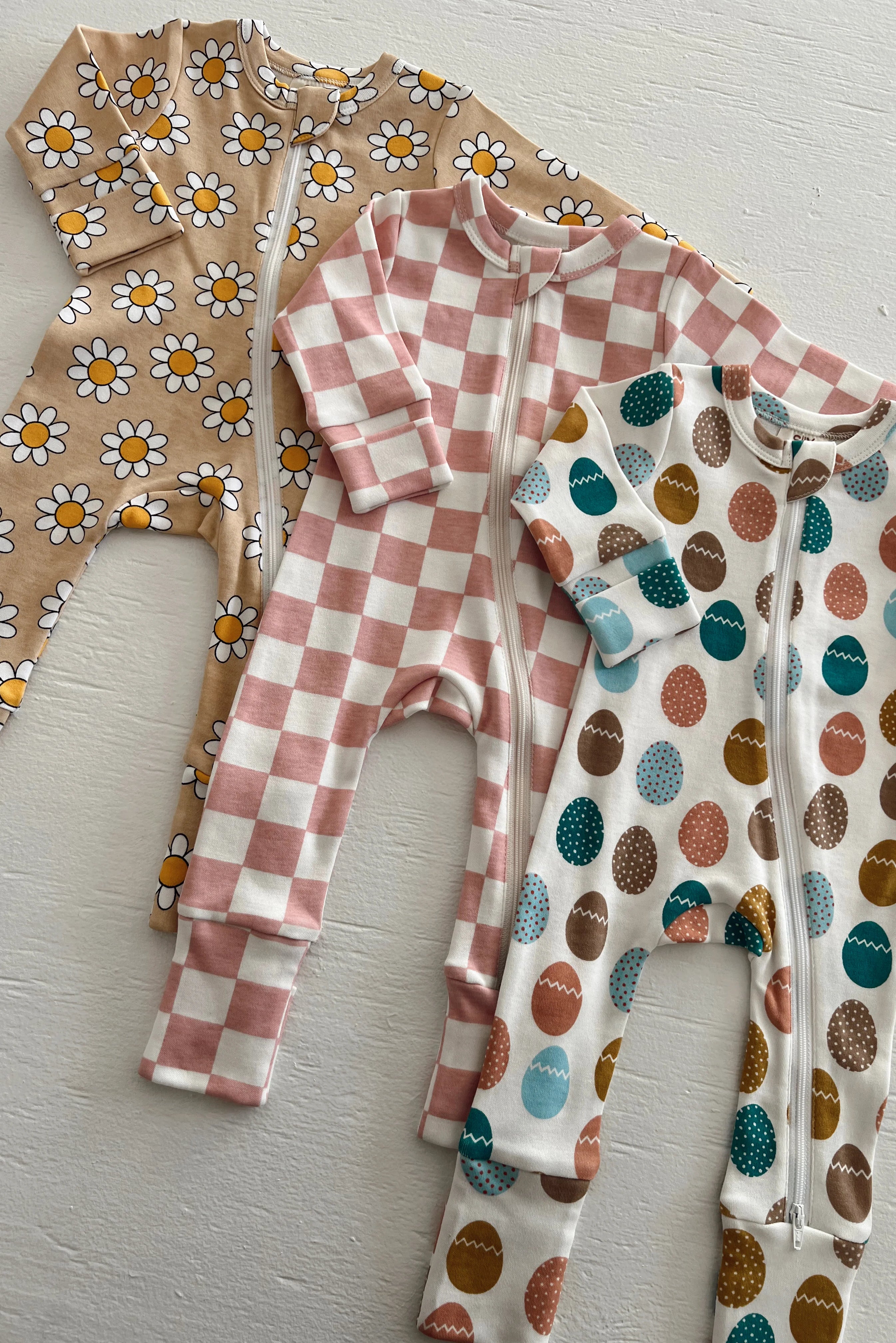 Three patterned baby onesies: floral, checkered, and egg designs, displayed on a light background.