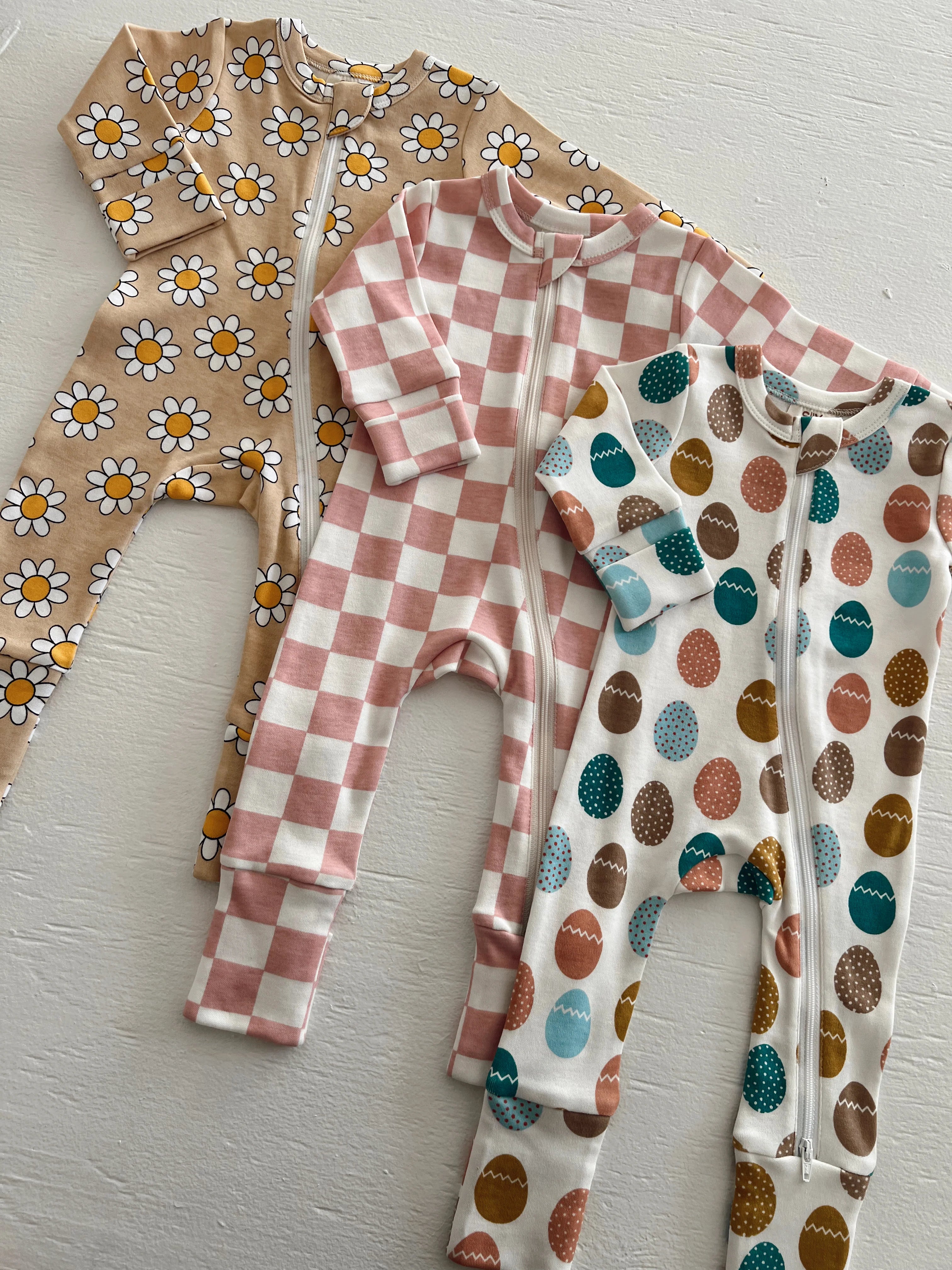 Three patterned baby onesies: floral, checkered, and egg designs, displayed on a light background.