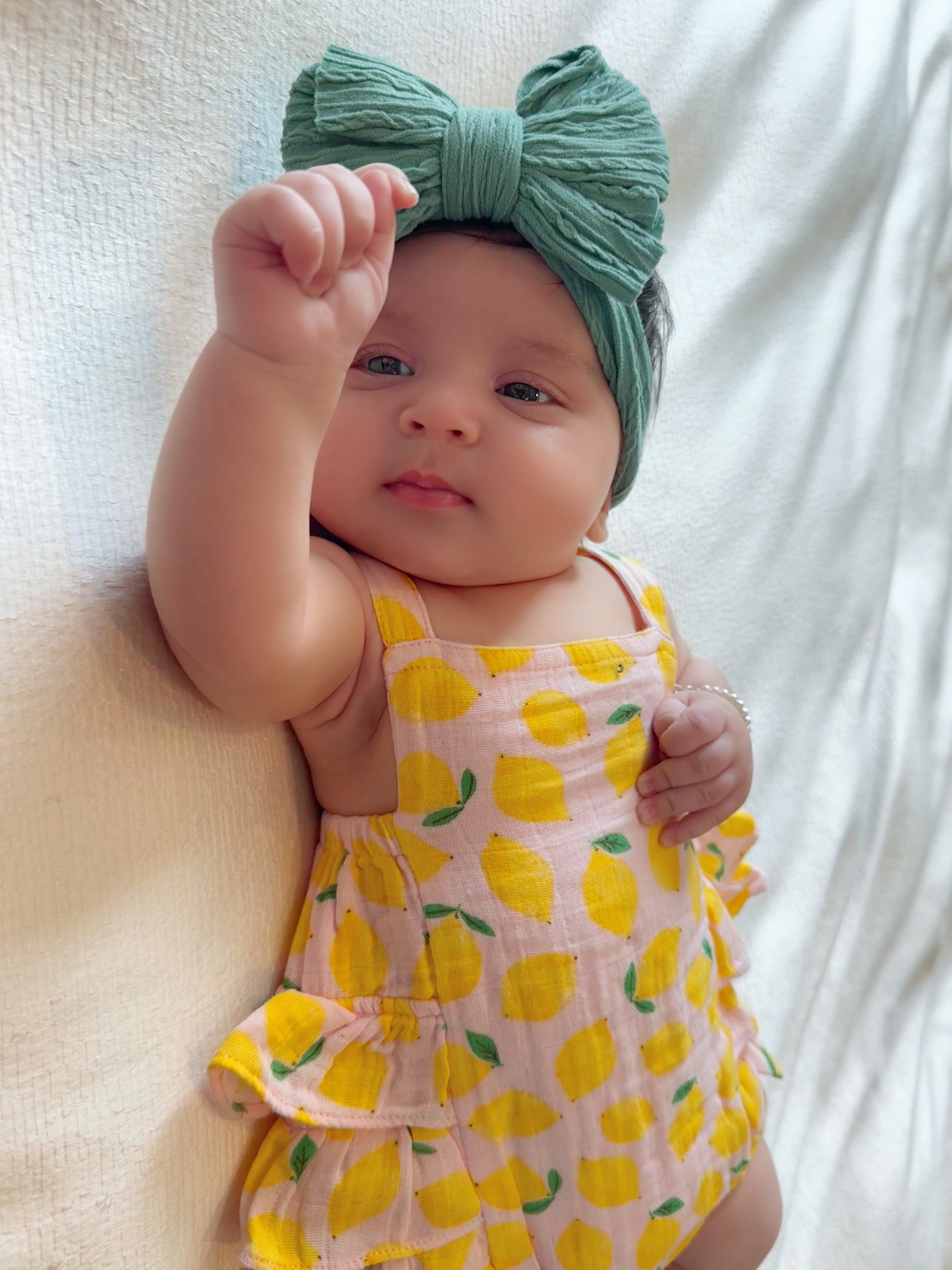 Baby girl in a yellow lemon dress and green bow headband, playfully raising her hand while sitting on a light-colored surface.