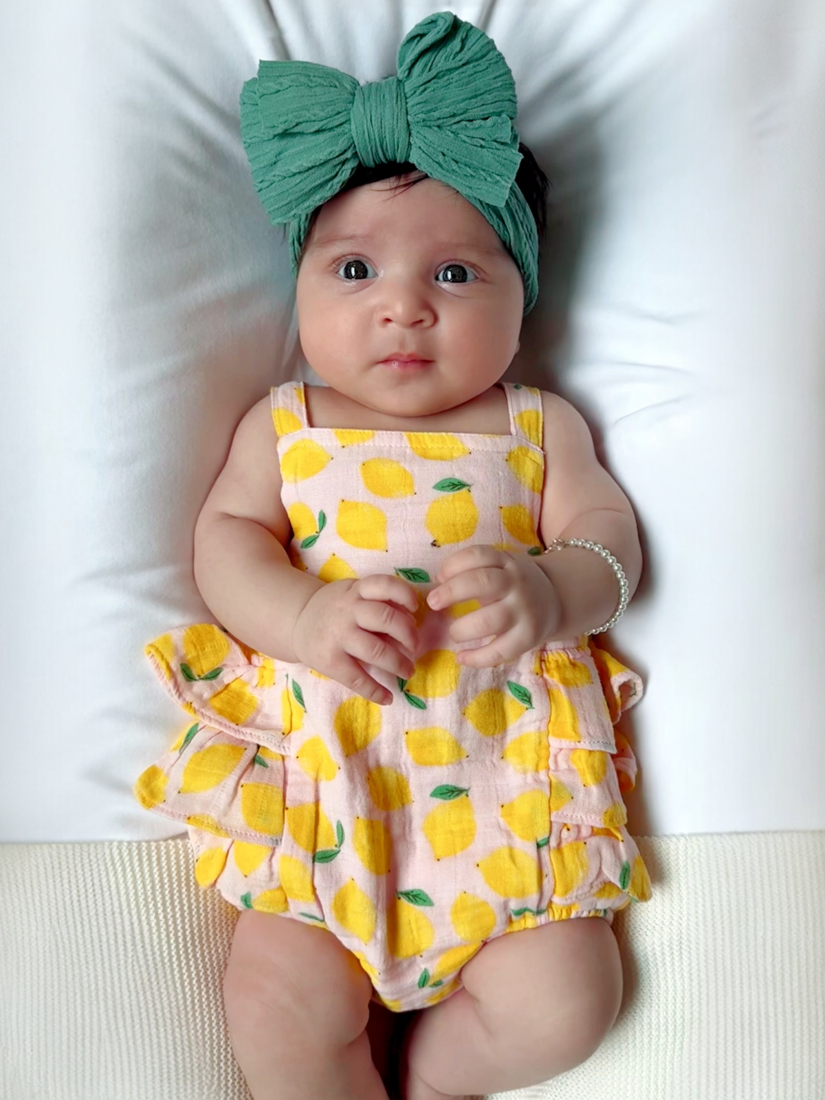 Baby in a lemon-patterned outfit and green bow, sitting on a white surface with a neutral background.