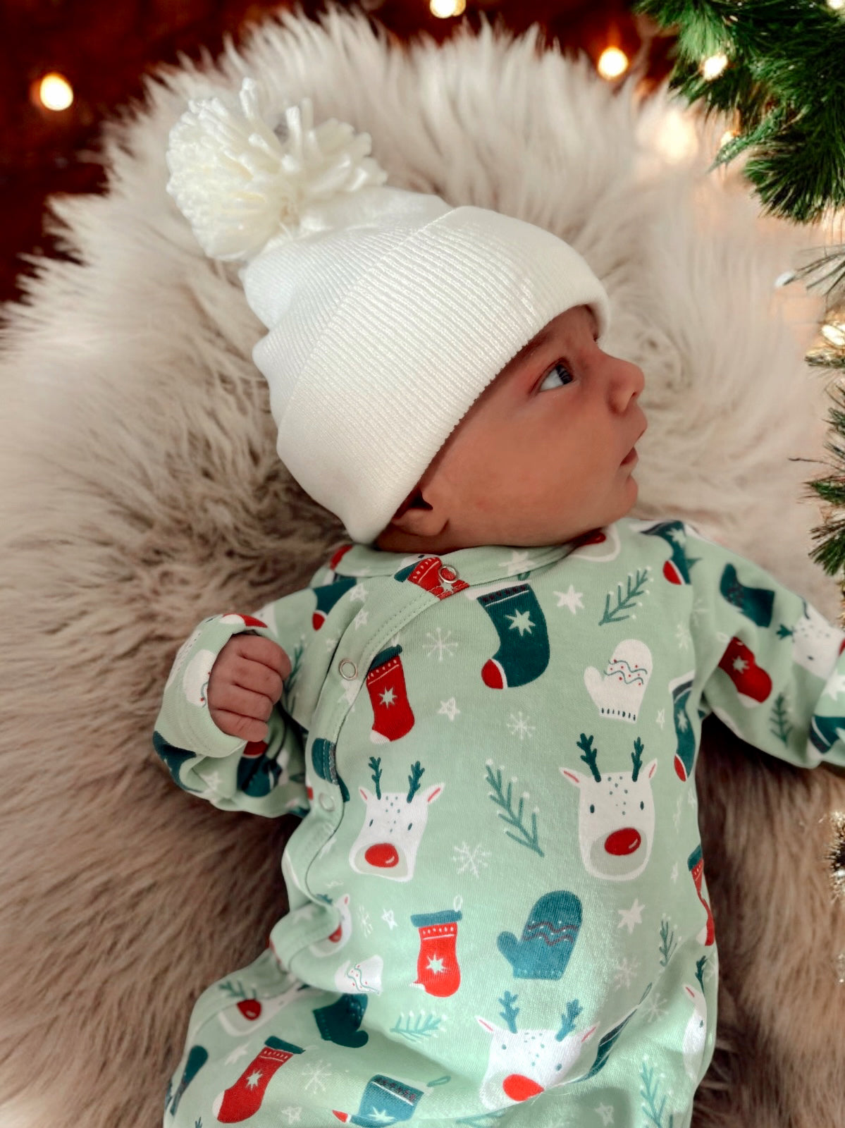 Infant in festive onesie with reindeer and mittens, wearing a white pom-pom beanie, resting on soft fur.