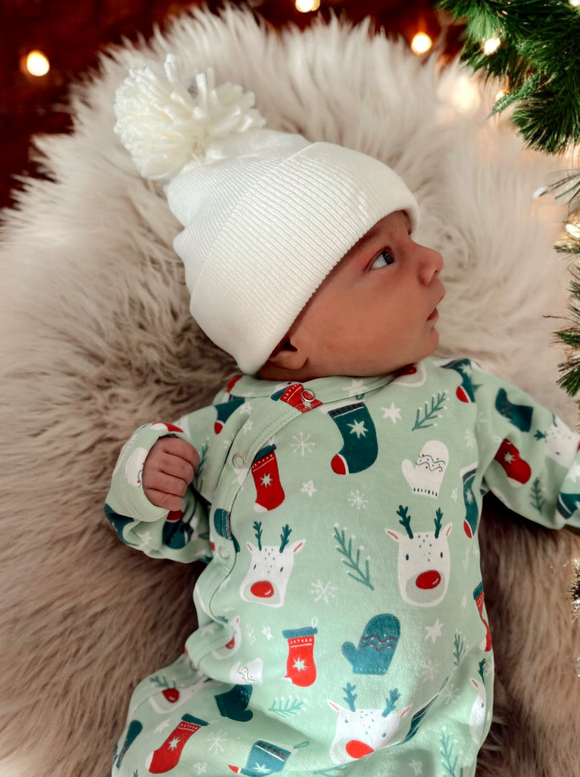 Infant in festive onesie with reindeer and mittens, wearing a white pom-pom beanie, resting on soft fur.