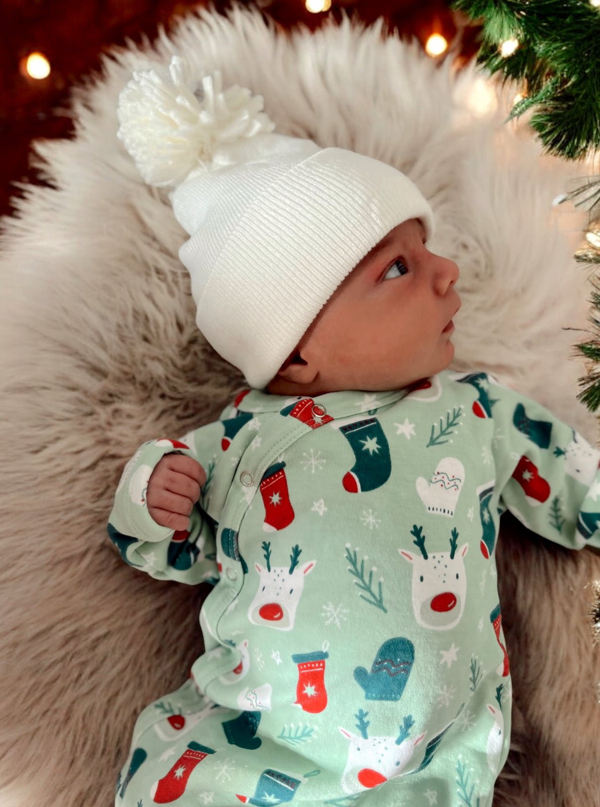 Infant in festive onesie with reindeer and mittens, wearing a white pom-pom beanie, resting on soft fur.