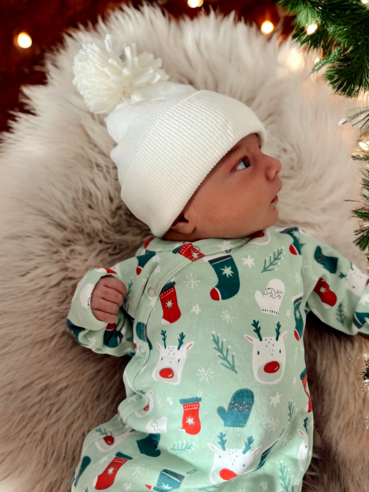 Infant in festive onesie with reindeer and mittens, wearing a white pom-pom beanie, resting on soft fur.