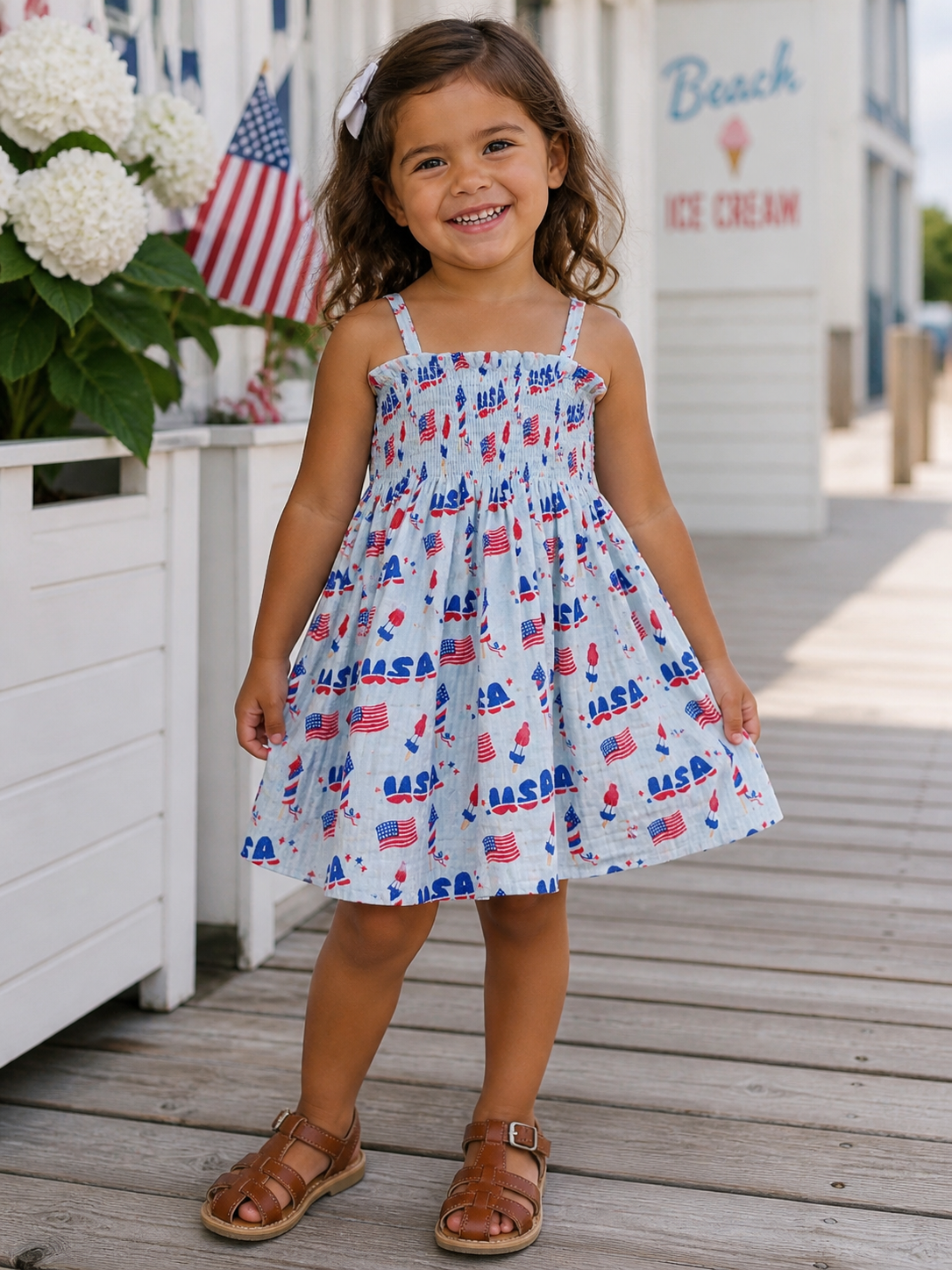 Smiling girl in blue dress with red and white designs, standing on wooden deck near beach and ice cream shop.