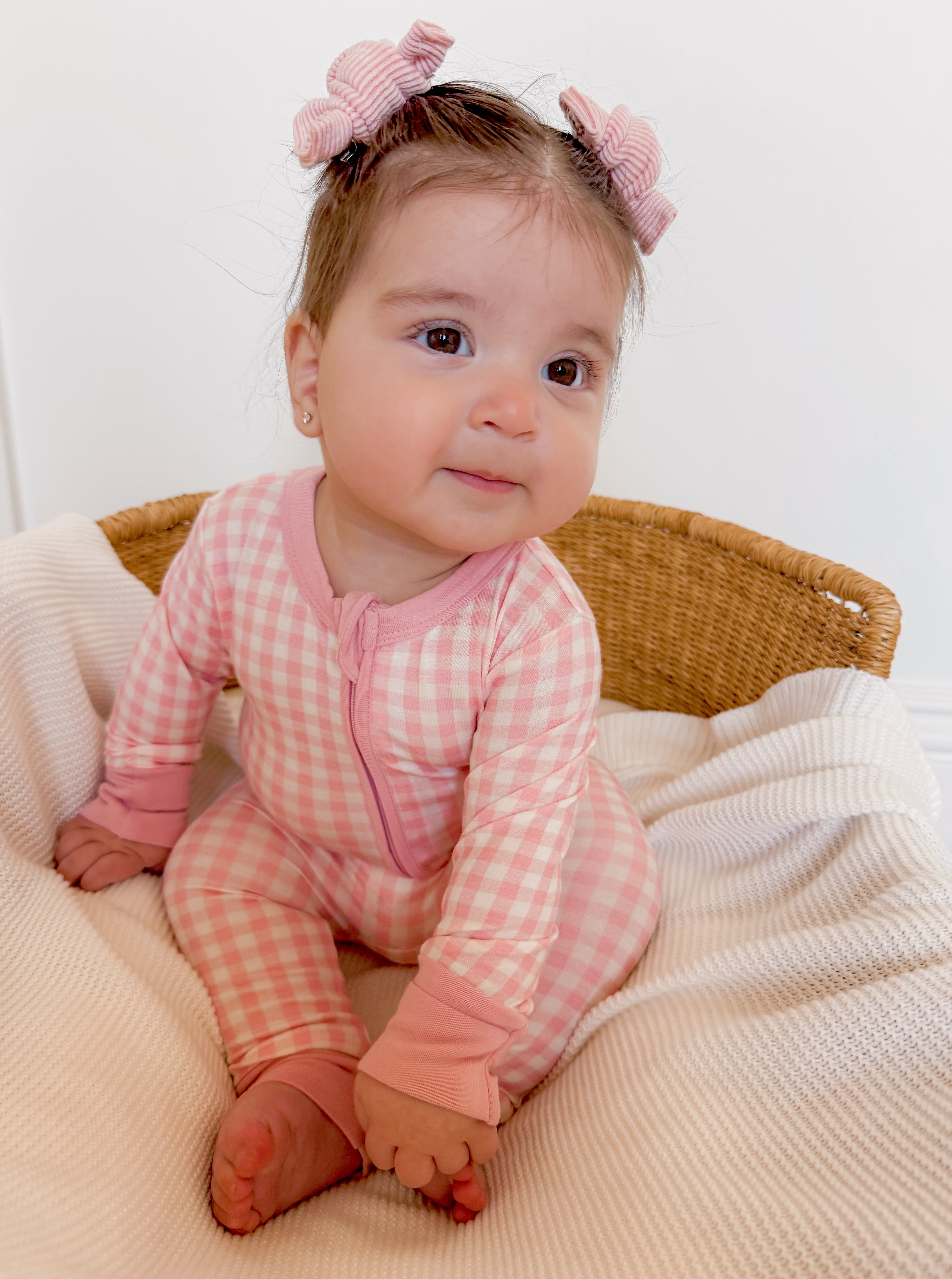 Baby girl in pink checkered pajamas with hair bows, sitting in a cozy basket on a soft blanket.