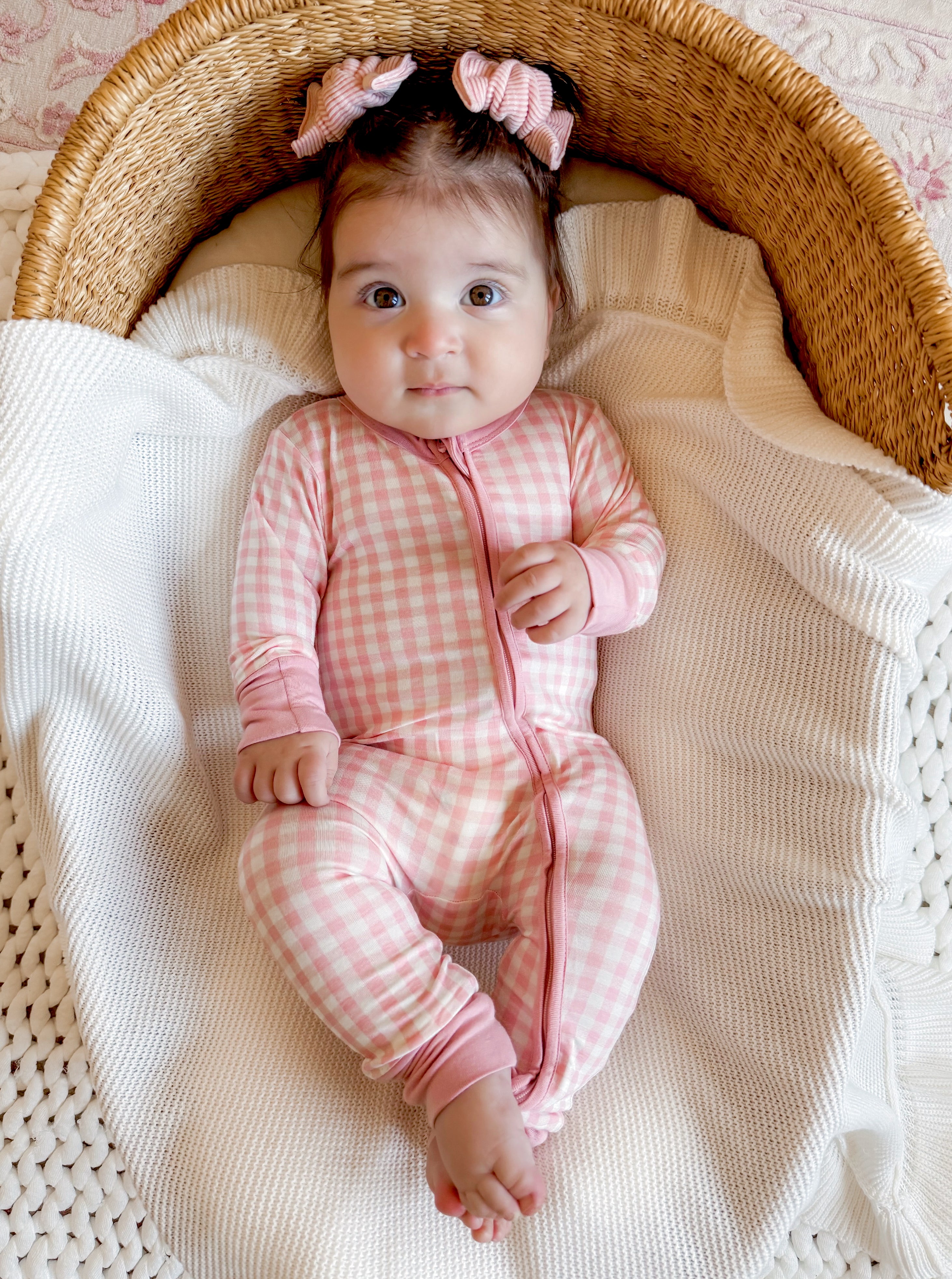 Baby in a pink checkered onesie sitting in a woven basket, surrounded by a cozy blanket.