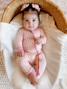 Baby in a pink checkered onesie sitting in a woven basket, surrounded by a cozy blanket.